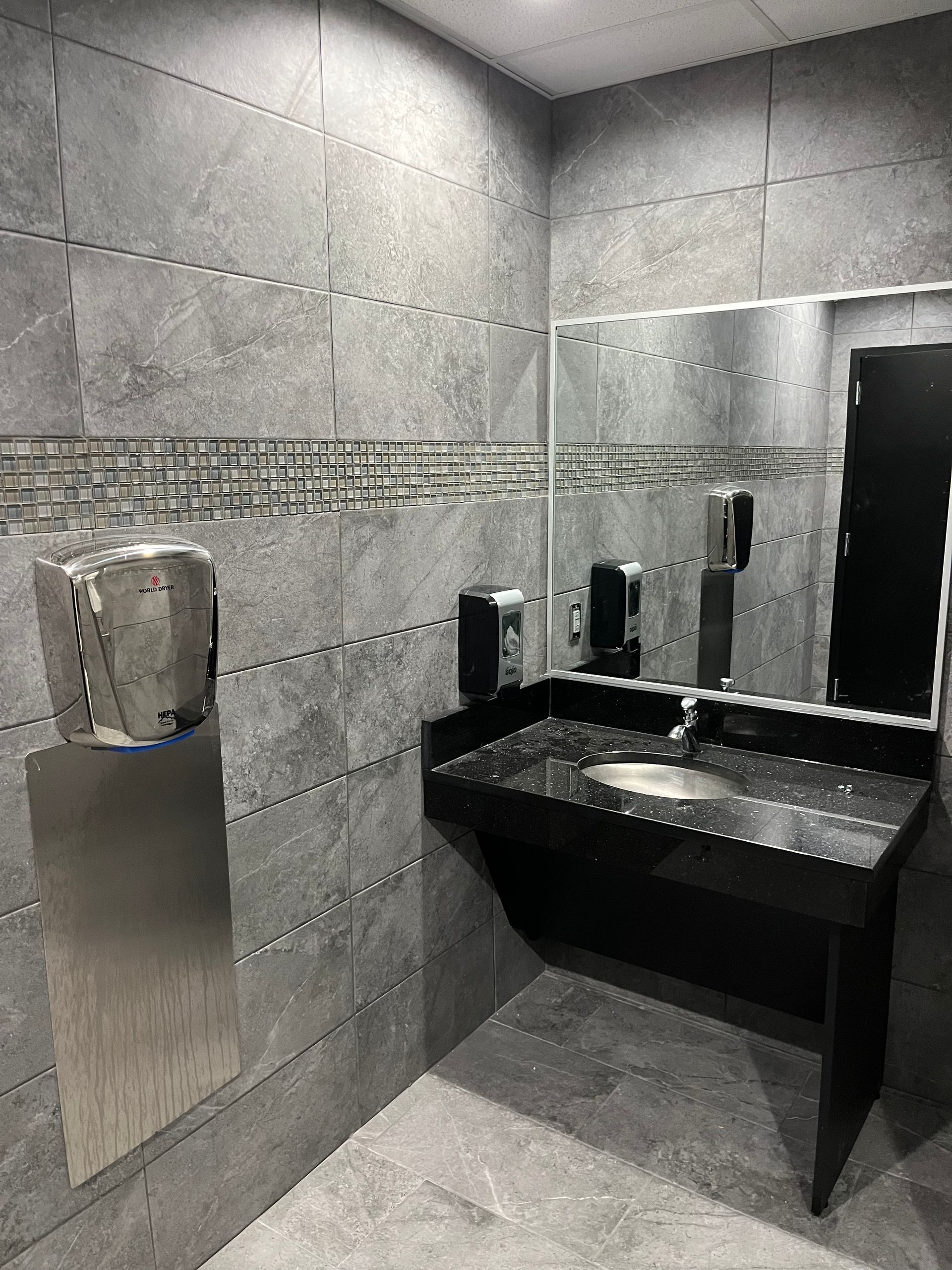 Modern gray-tiled restroom with a black sink counter and mirror, plus a wall-mounted soap dispenser.
