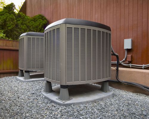 Two outdoor air conditioning units on gravel next to a wooden fence.