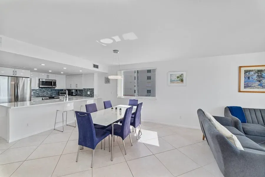 Bright, open-plan dining and kitchen space. White walls, tile floor, blue upholstered chairs, and a gray sofa.