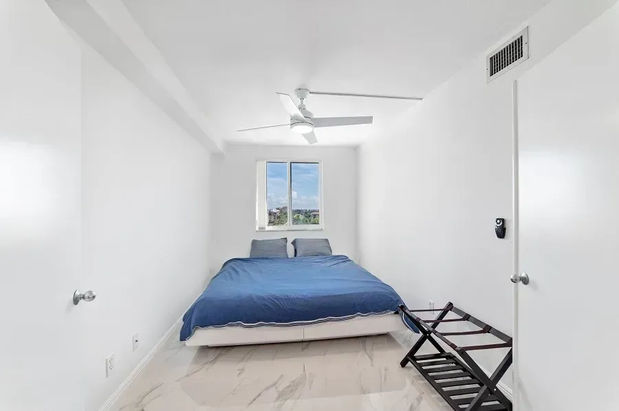 White bedroom with a bed, luggage rack, and window; ceiling fan.