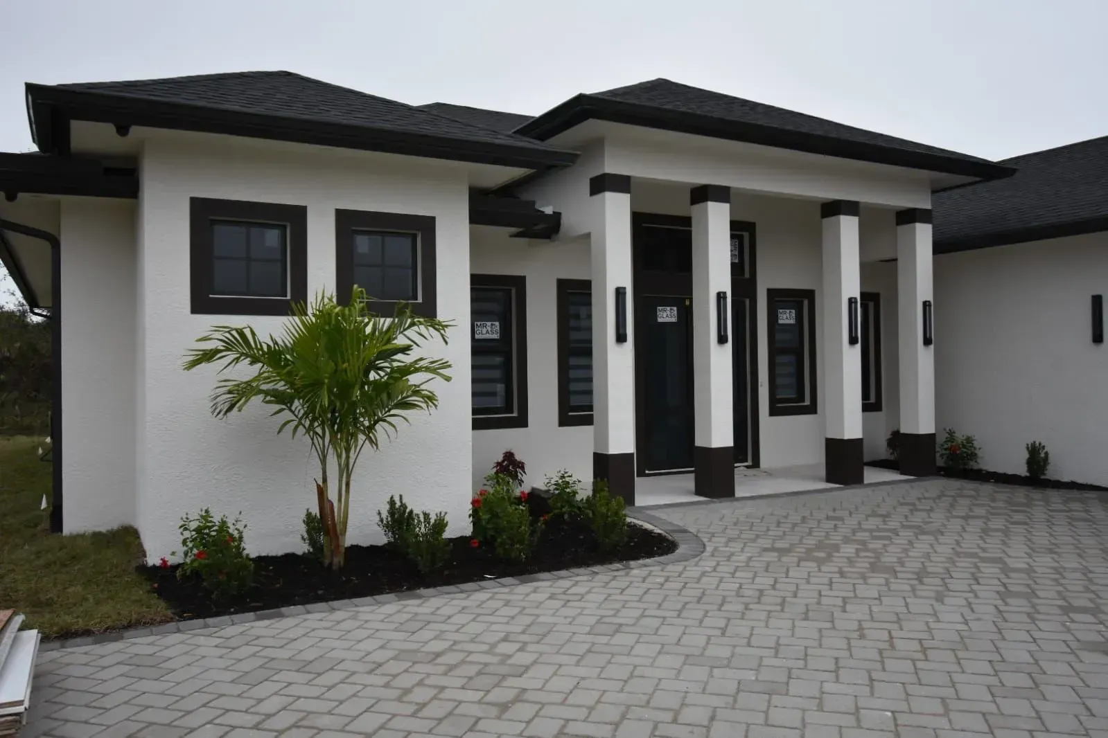 White house with black trim, columns, and a gray brick driveway. Small palm tree and bushes in front.