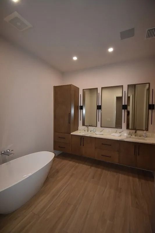 Modern bathroom with wood cabinetry, three mirrors, a freestanding tub, and wood-look tile flooring.