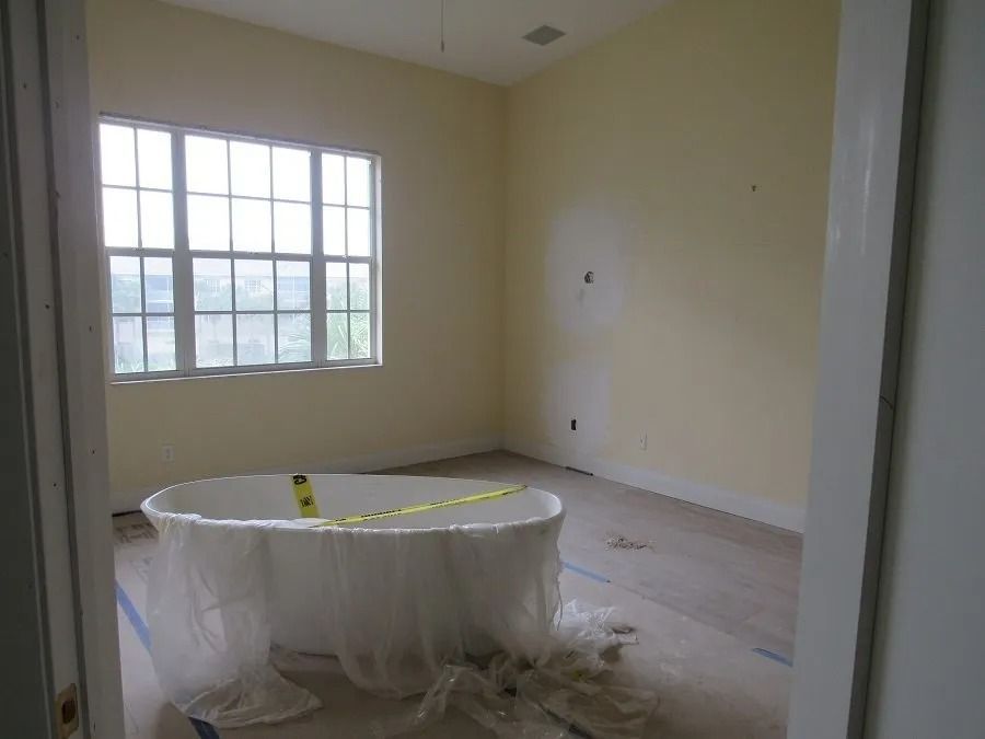 Bathroom with a large white bathtub covered in plastic, by a window. Pale yellow walls and unfinished floor.