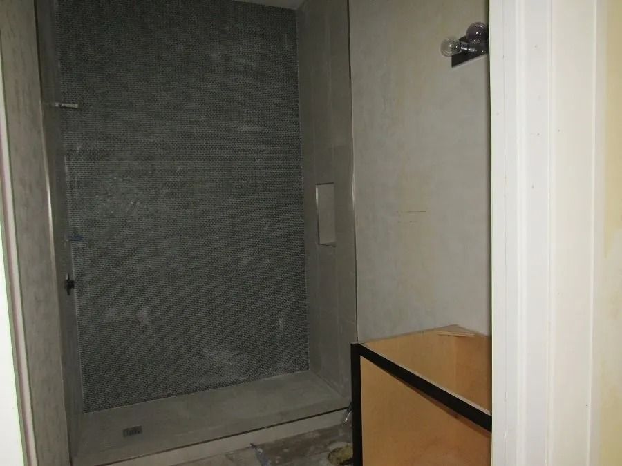 A mostly-finished shower with gray tile, a glass door, and unfinished walls. A light-colored vanity cabinet is visible.