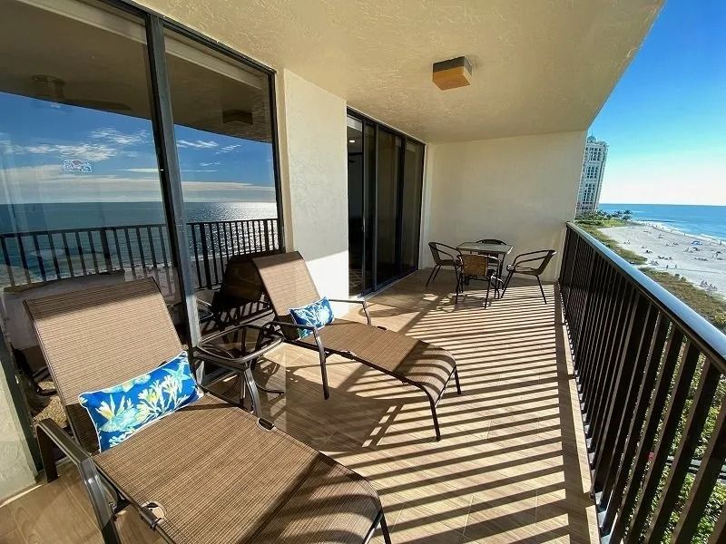 Balcony with beach view: lounge chairs, table, chairs, dark railing, ocean, and bright blue sky.