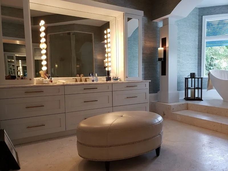 Spacious white bathroom with vanity lights, an oval ottoman, and a soaking tub near a window.