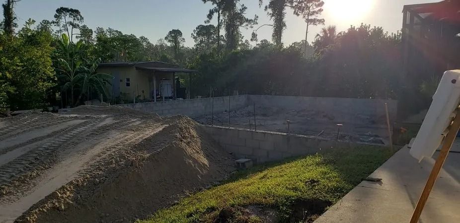 A small house, dirt pile, and concrete construction area are in a wooded setting with sunlight.