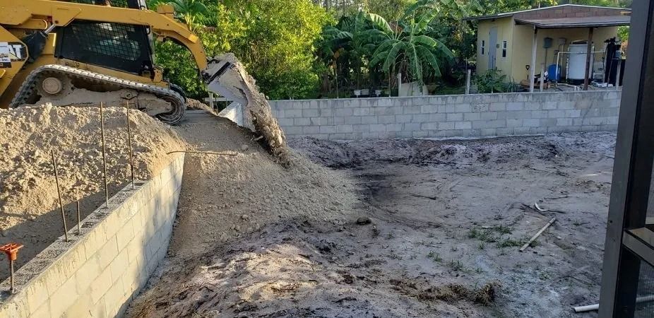 A yellow bulldozer dumping sand into a construction site. A house and trees are in the background.