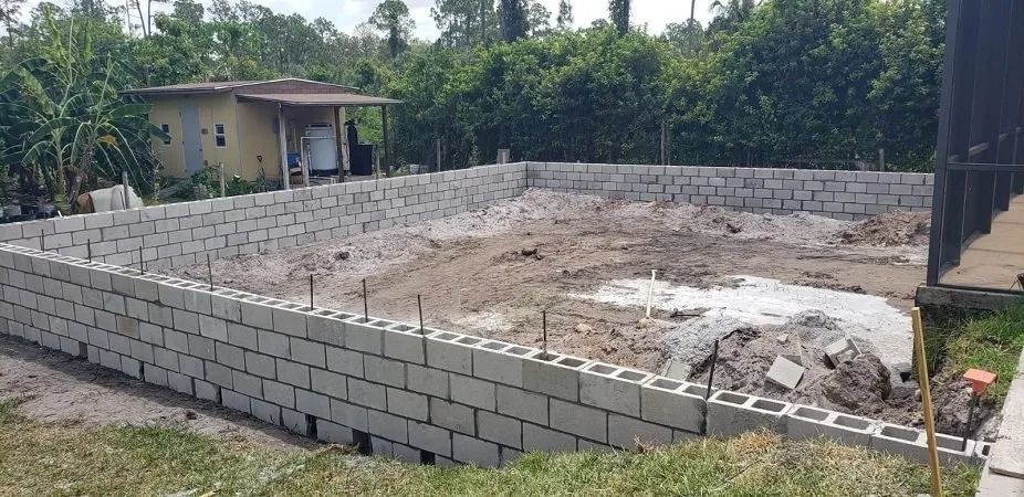 A rectangular concrete block structure under construction, set in a yard with a small building in the background.