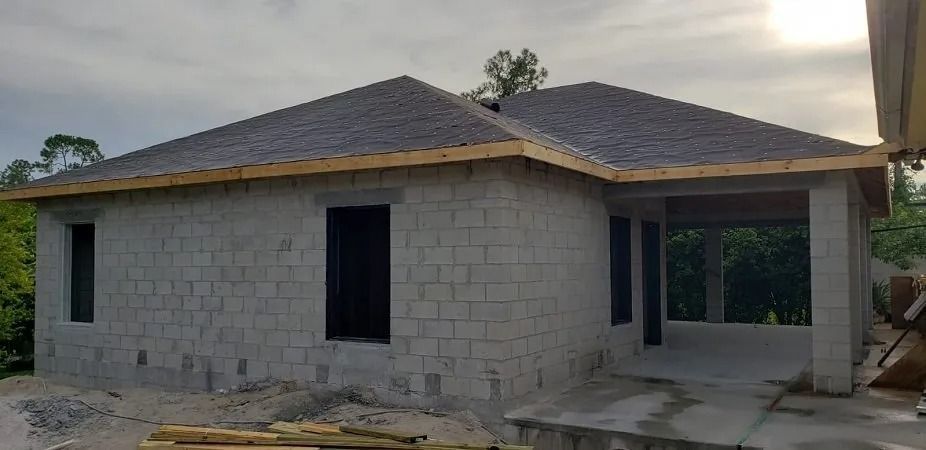 A gray cinder block house under construction, with a dark roof and open windows.