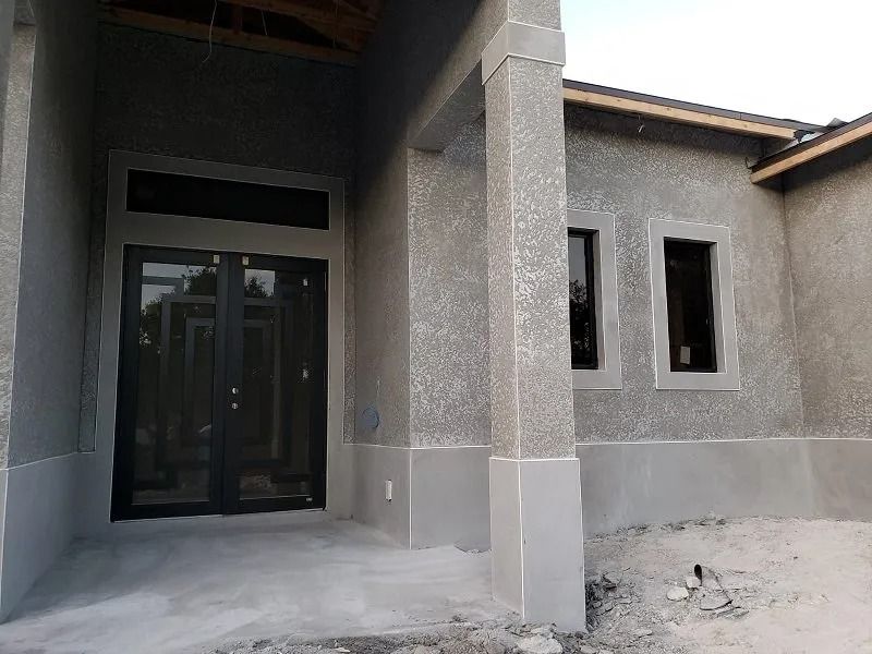 A gray stucco house under construction, with a dark double door entry and two dark framed windows.