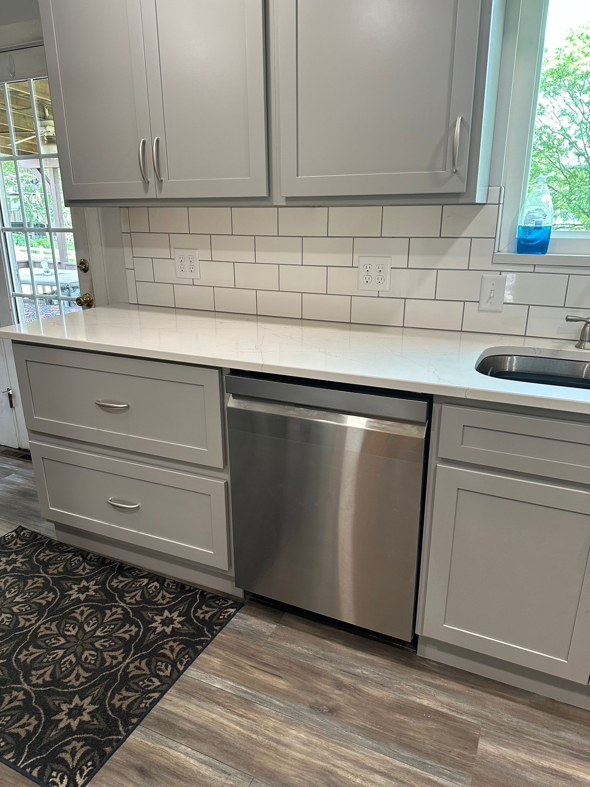 Modern kitchen with gray cabinets, stainless dishwasher, white tile backsplash, and wood-look floor.