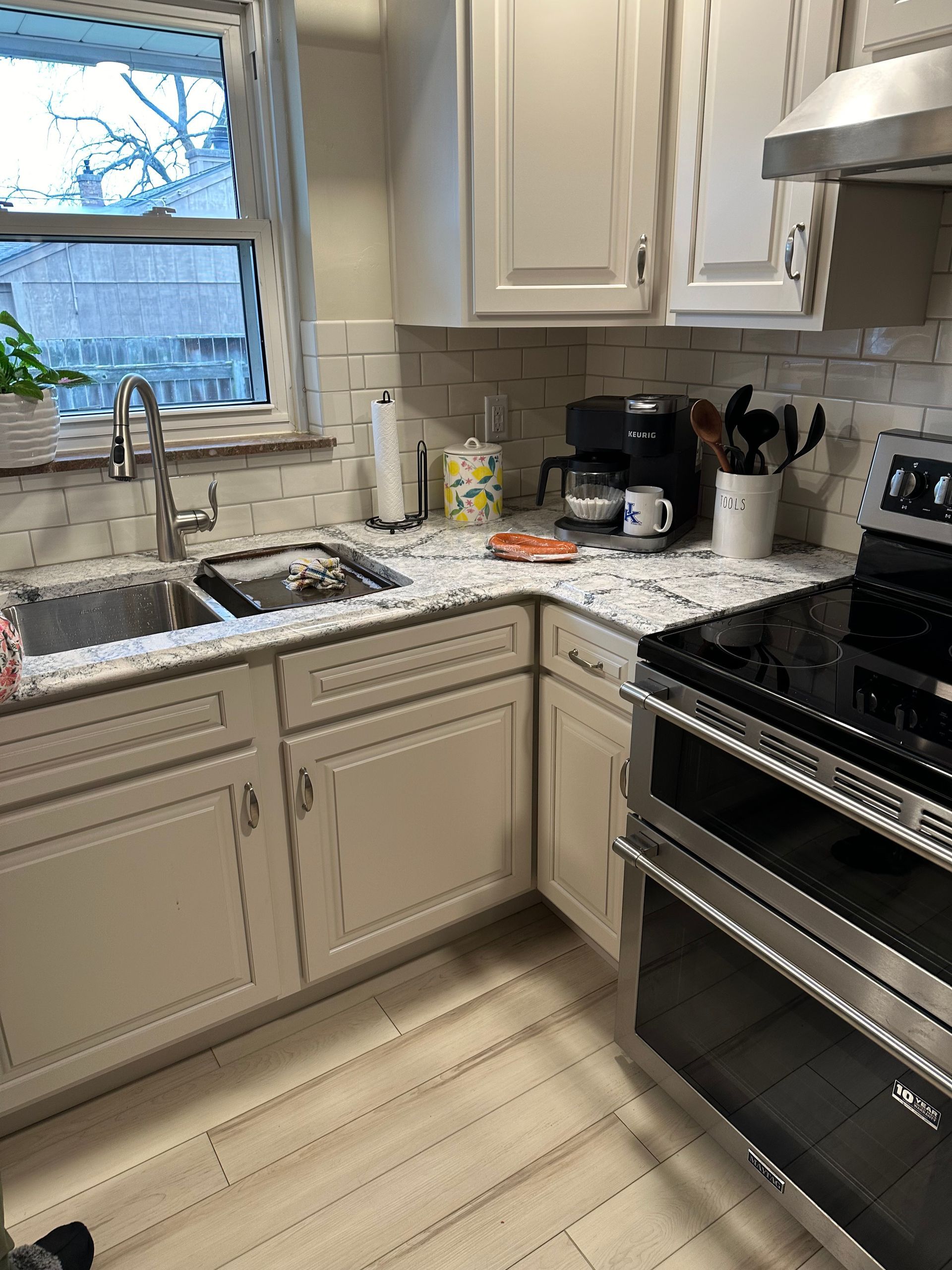 Kitchen corner with beige cabinets, stainless steel stove, sink by a window, and coffee maker on the counter.