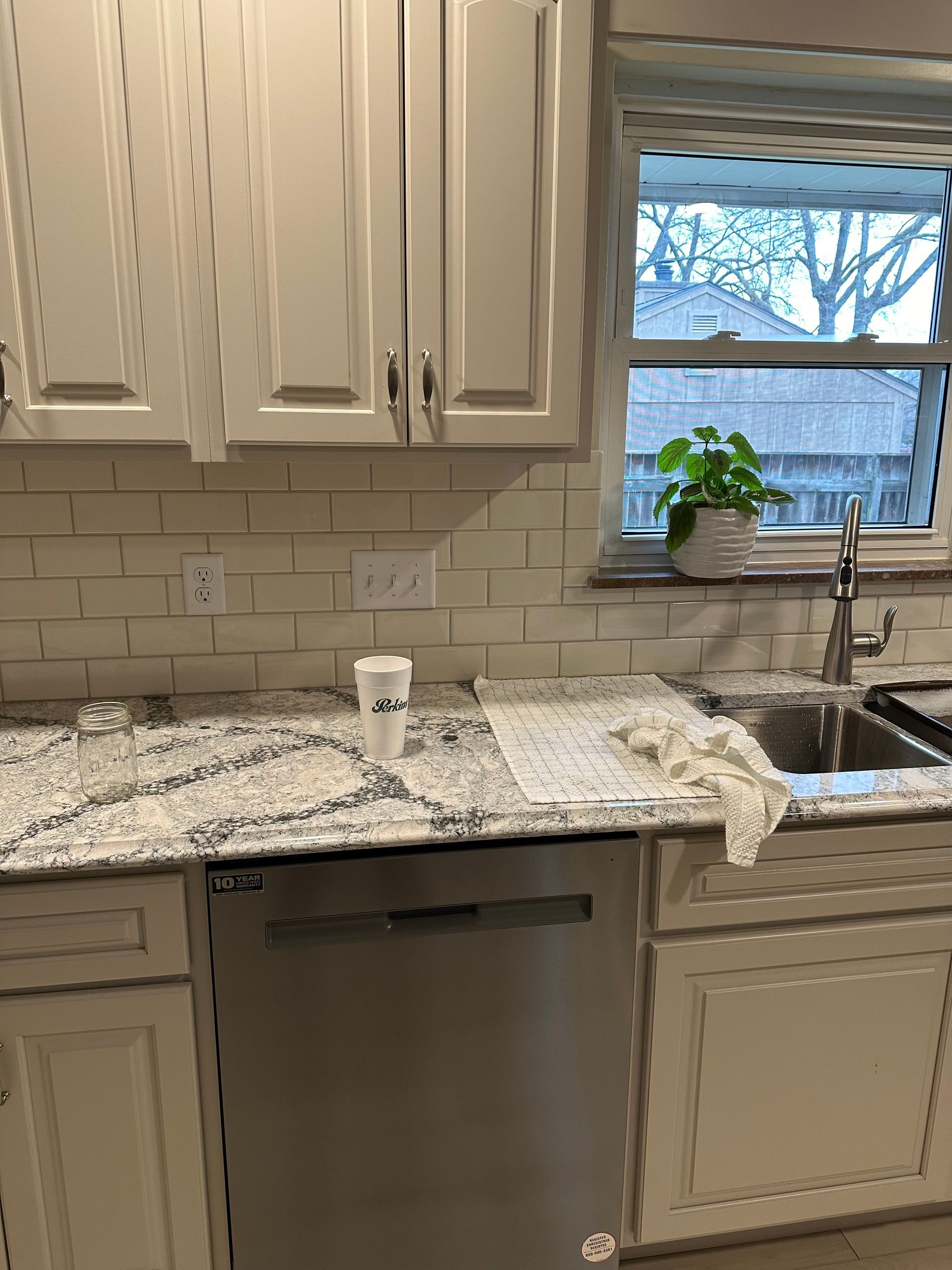 Modern kitchen with white cabinets, speckled granite counter, stainless dishwasher, sink, and window with plant.