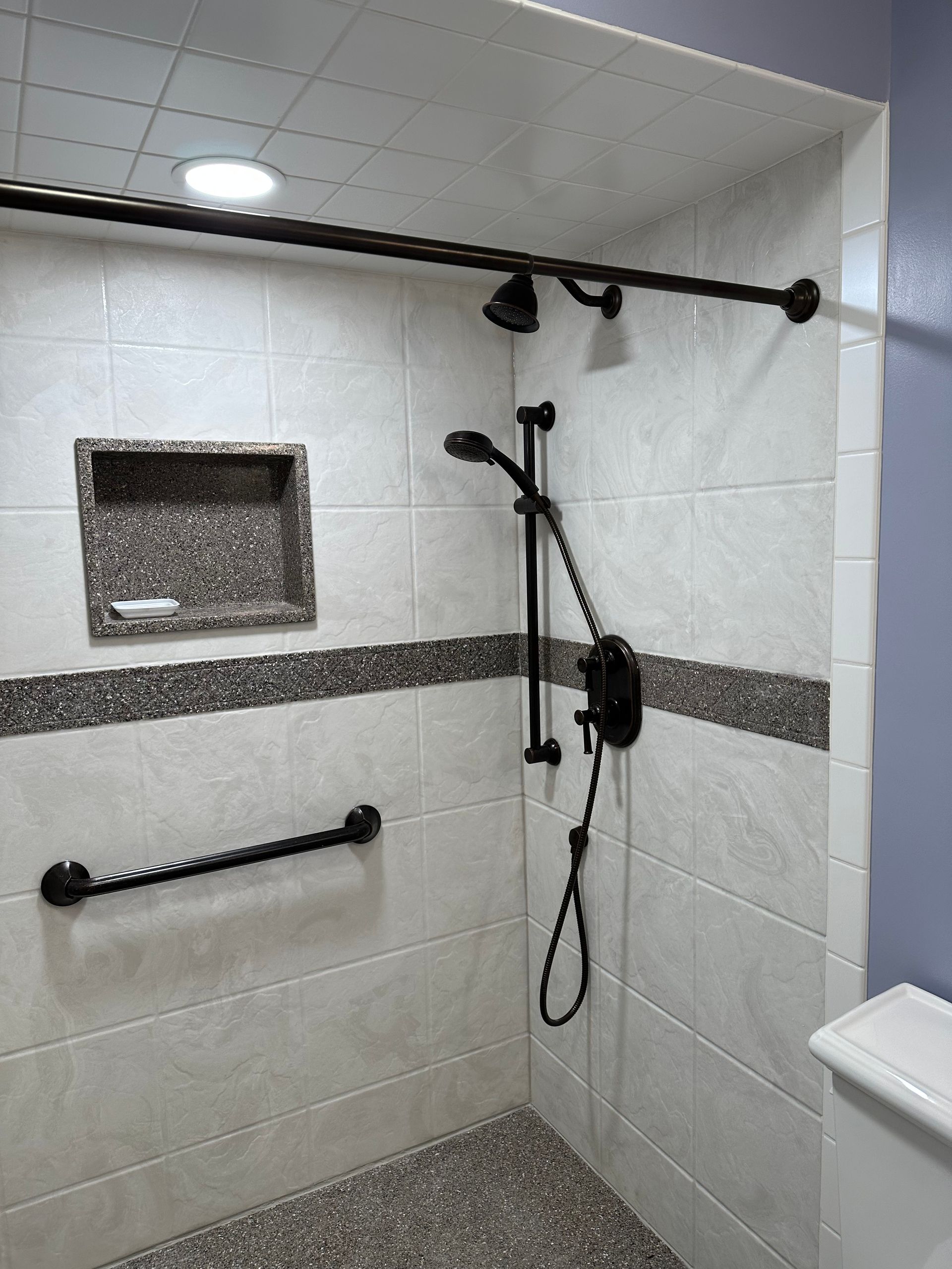 Accessible shower with grab bars, handheld showerhead, and a recessed shelf in a tiled bathroom.