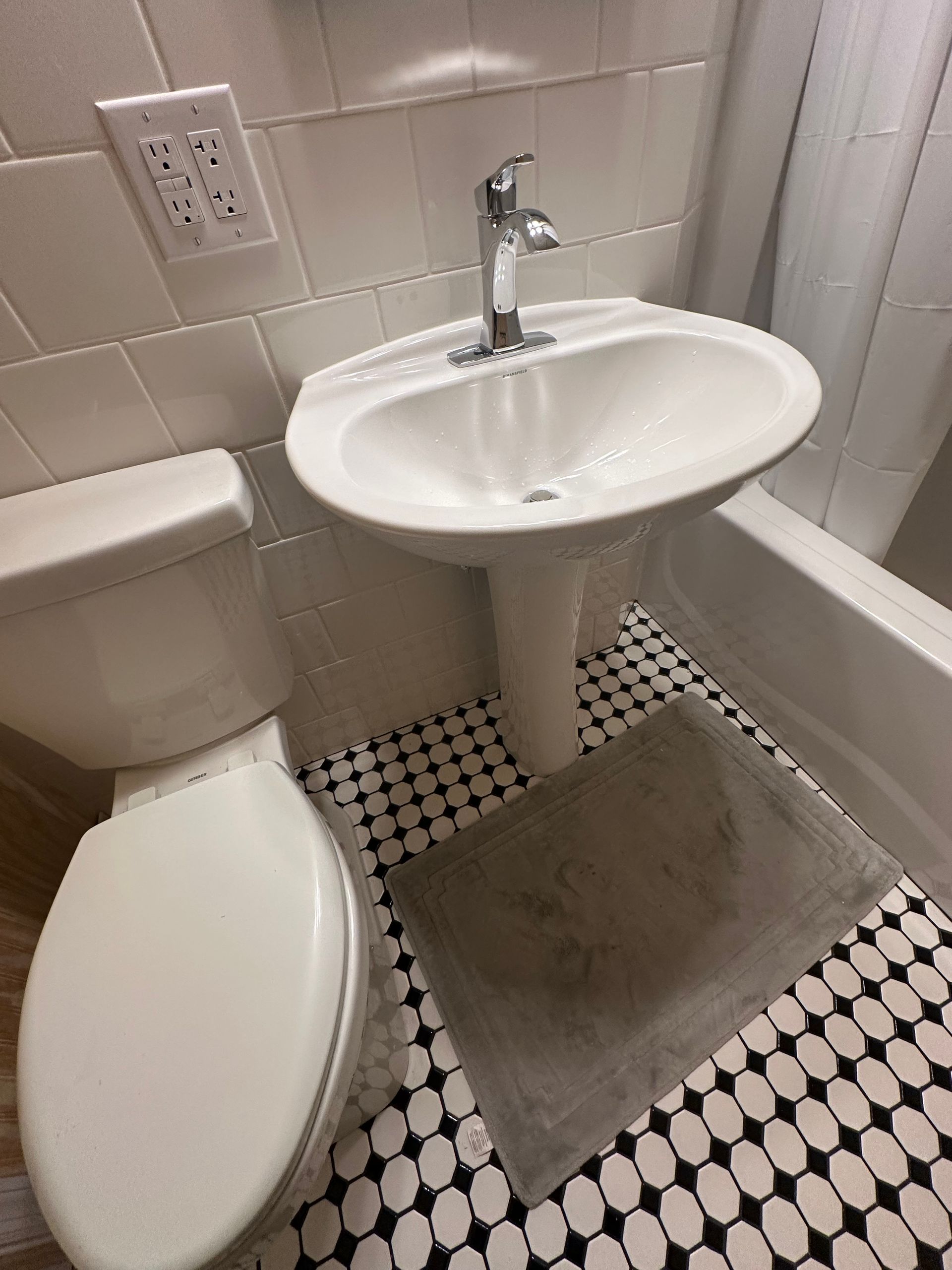 Small bathroom with toilet, pedestal sink, patterned tile floor, and gray bath mat.