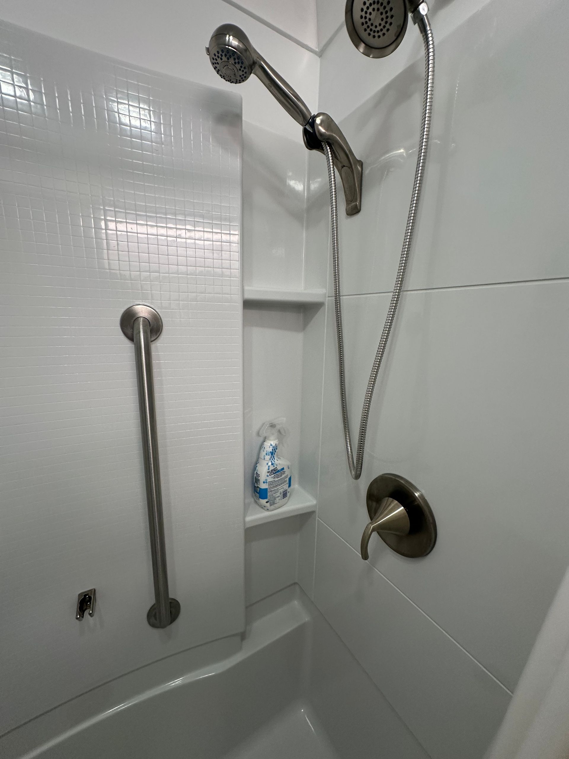 White tiled shower with grab bars, built-in shelf, handheld showerhead, and a bathtub edge.