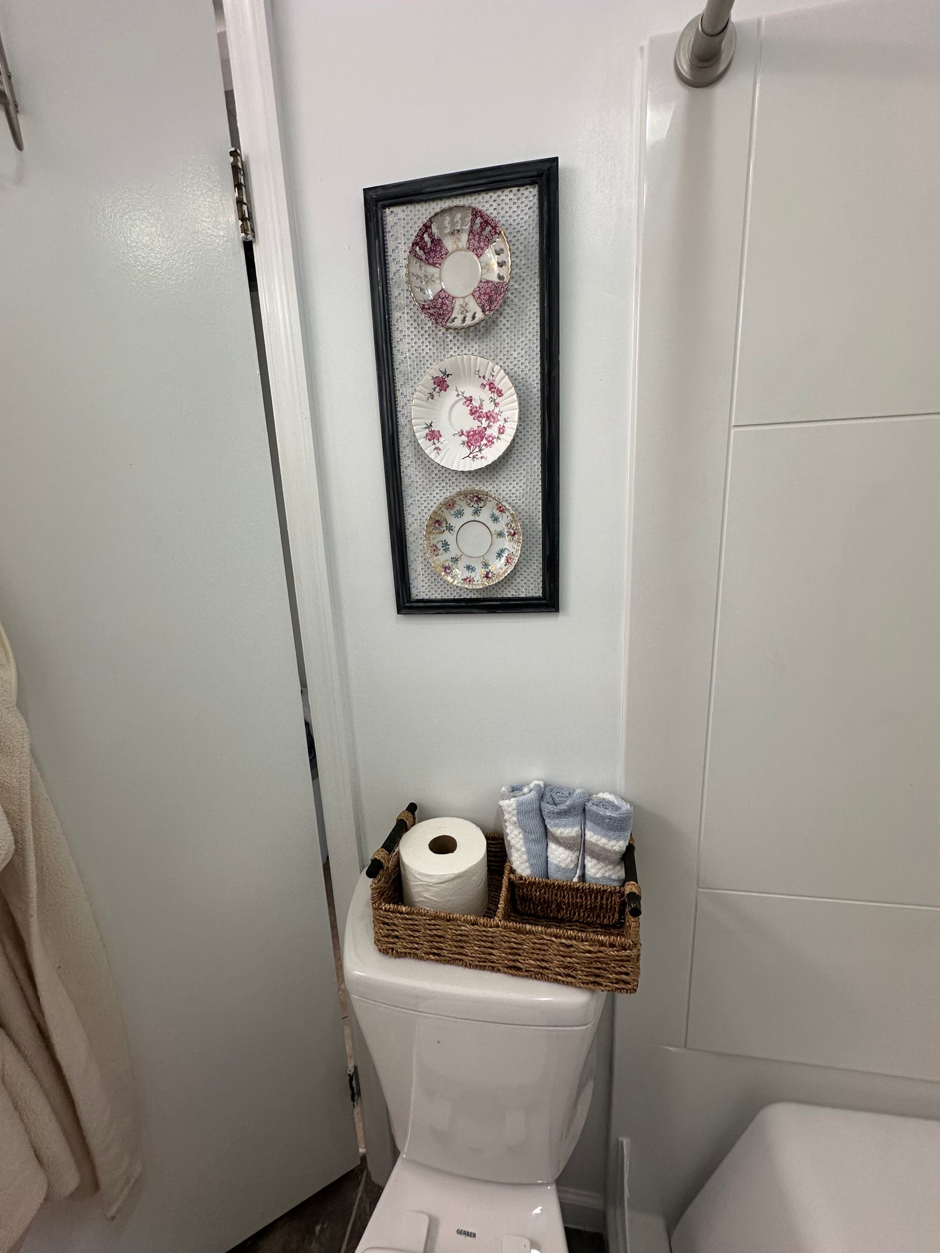 Small bathroom toilet with wicker tray holding toilet paper and wipes, framed floral wall art above.