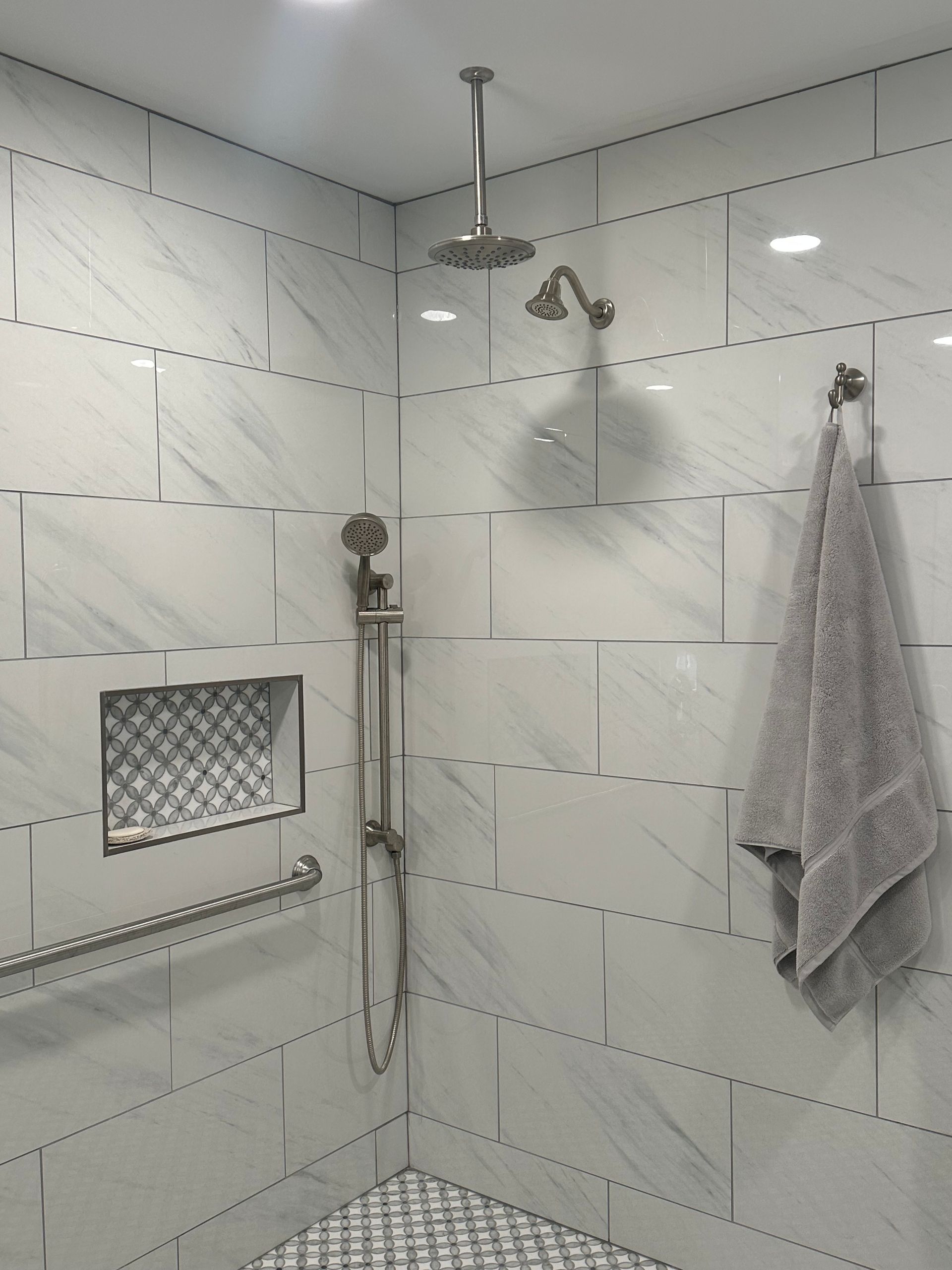 Modern tiled bathroom shower with wall-mounted showerhead, handheld sprayer, towel, and built-in niche.