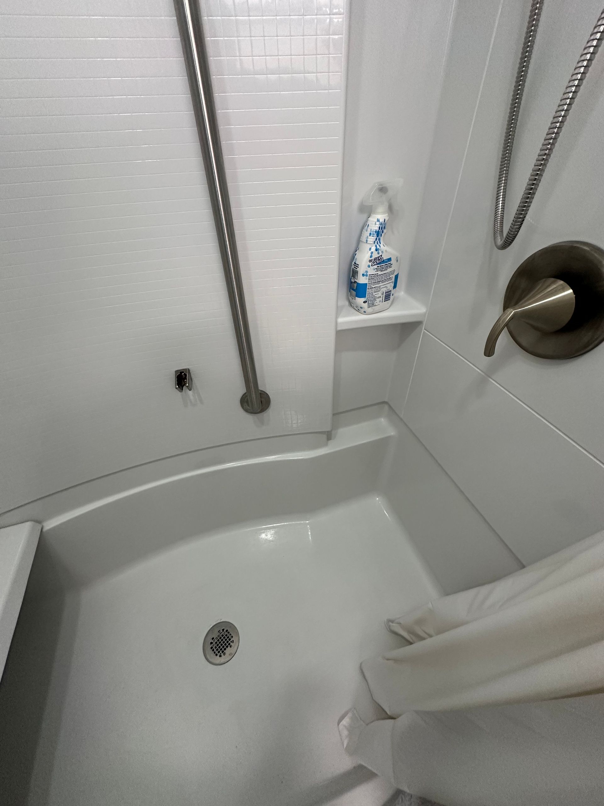 Small white shower with grab bars, bench seat, handheld shower head, and curtain in a bathroom
