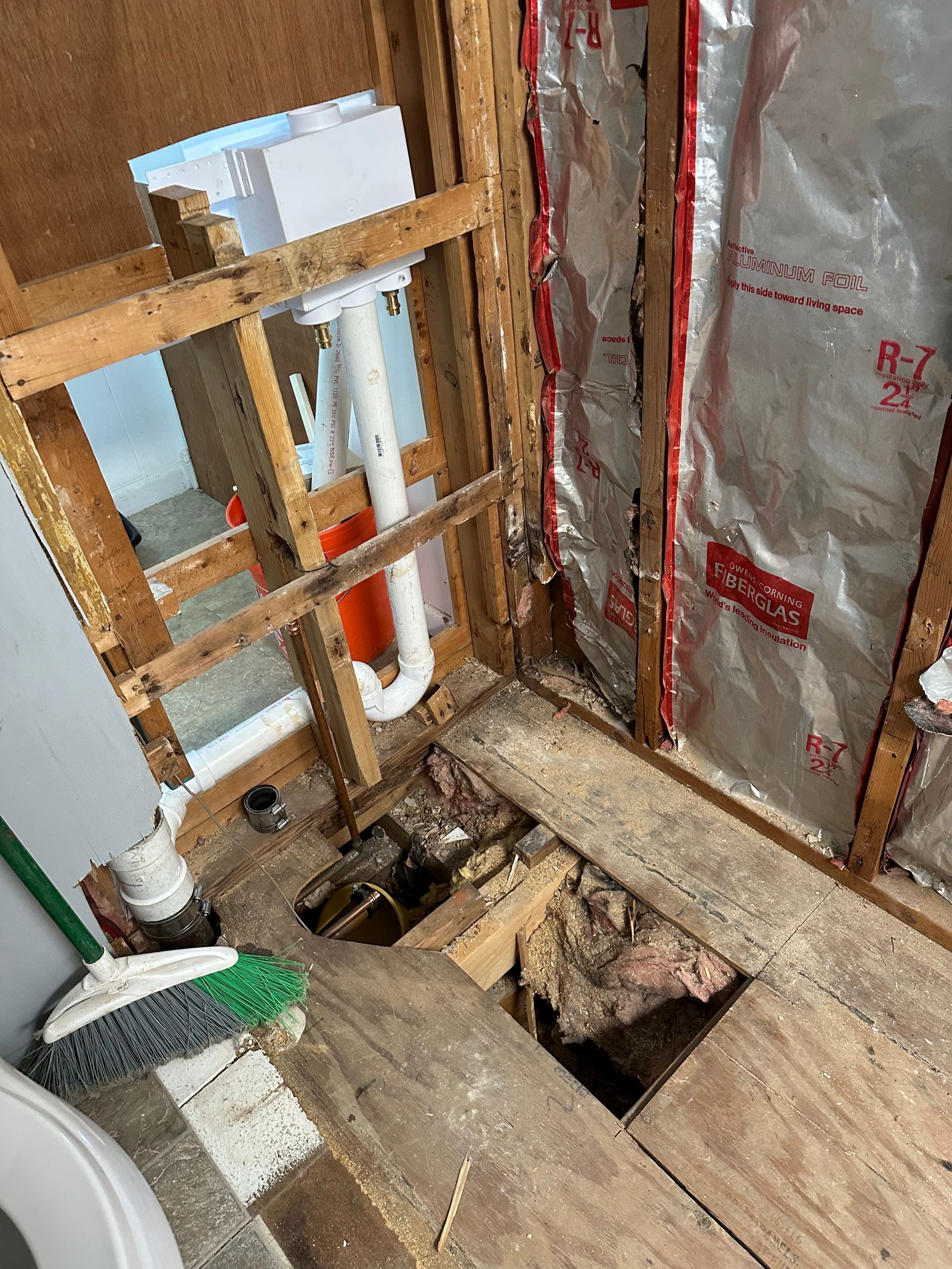 Bathroom floor cut open near toilet, exposing plumbing and joists, with insulation and wood framing visible.