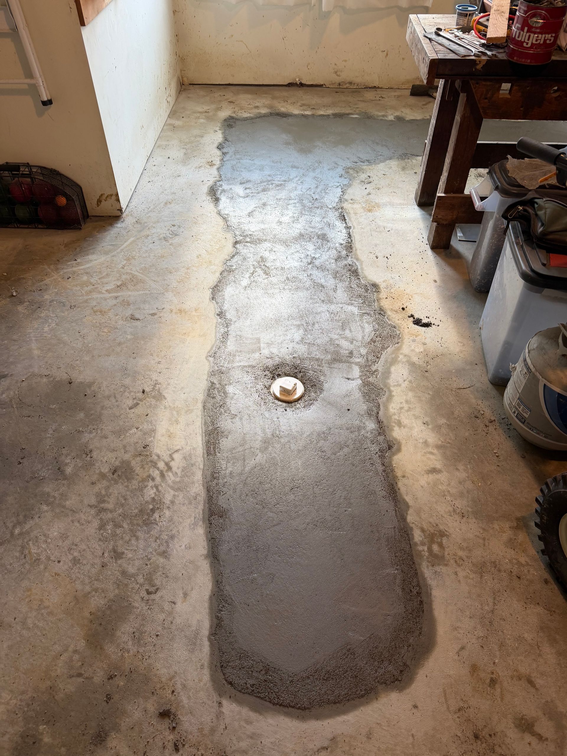 Narrow gray concrete patch on a tan floor in a cluttered room, with a small drain near the center.