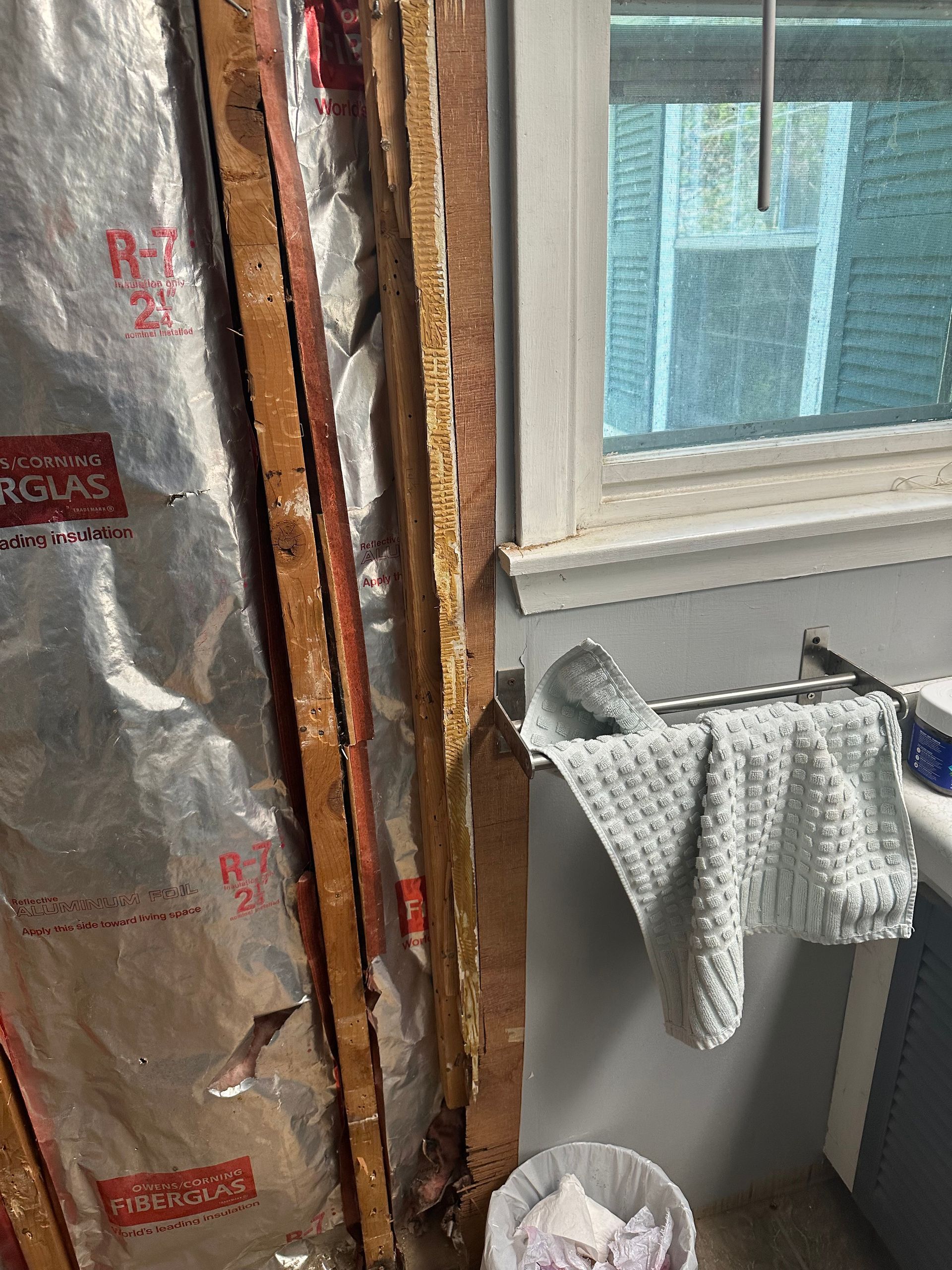 Interior wall opening with exposed insulation beside a window and hanging gloves over a radiator.