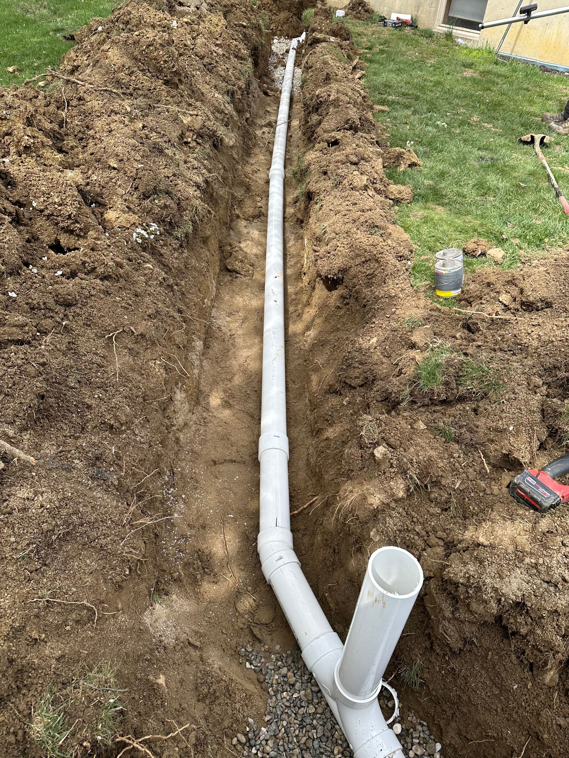 White PVC pipe laid in a narrow trench beside a grassy yard, with an elbow joint and vertical pipe opening.