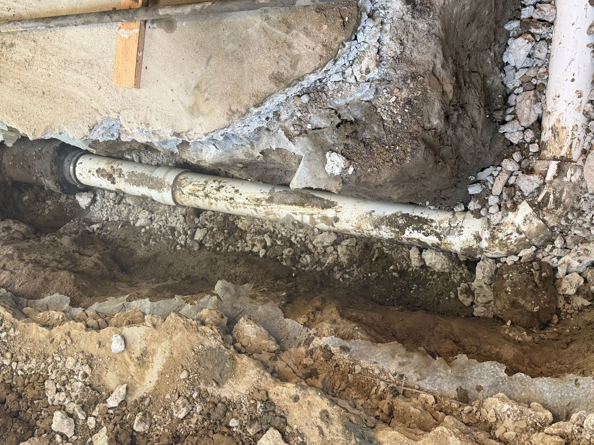 Exposed white pipe running through a dirt trench beside a stone wall and concrete foundation