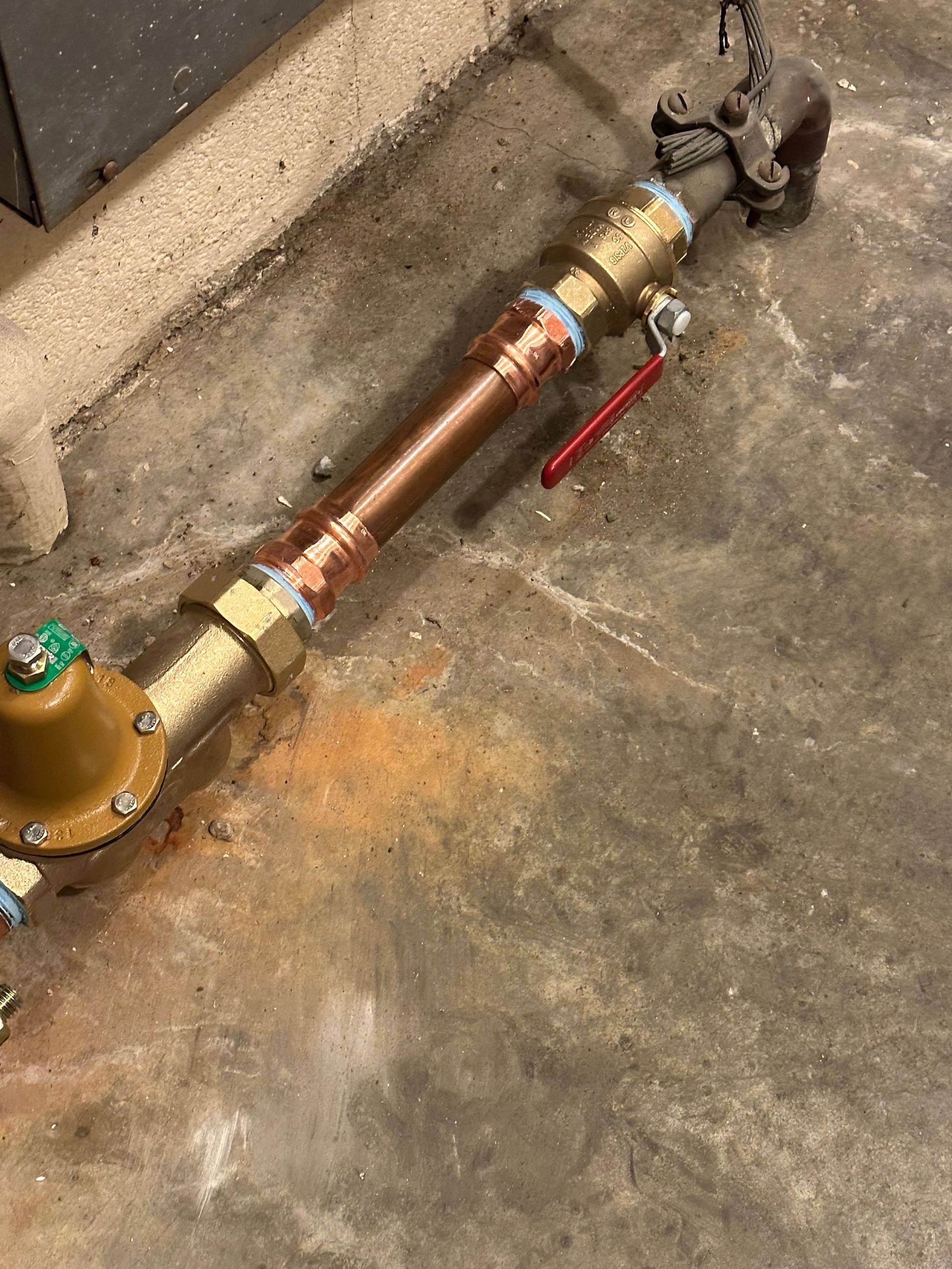 Copper pipe assembly with brass valves and a red lever on a concrete floor beside a wall.