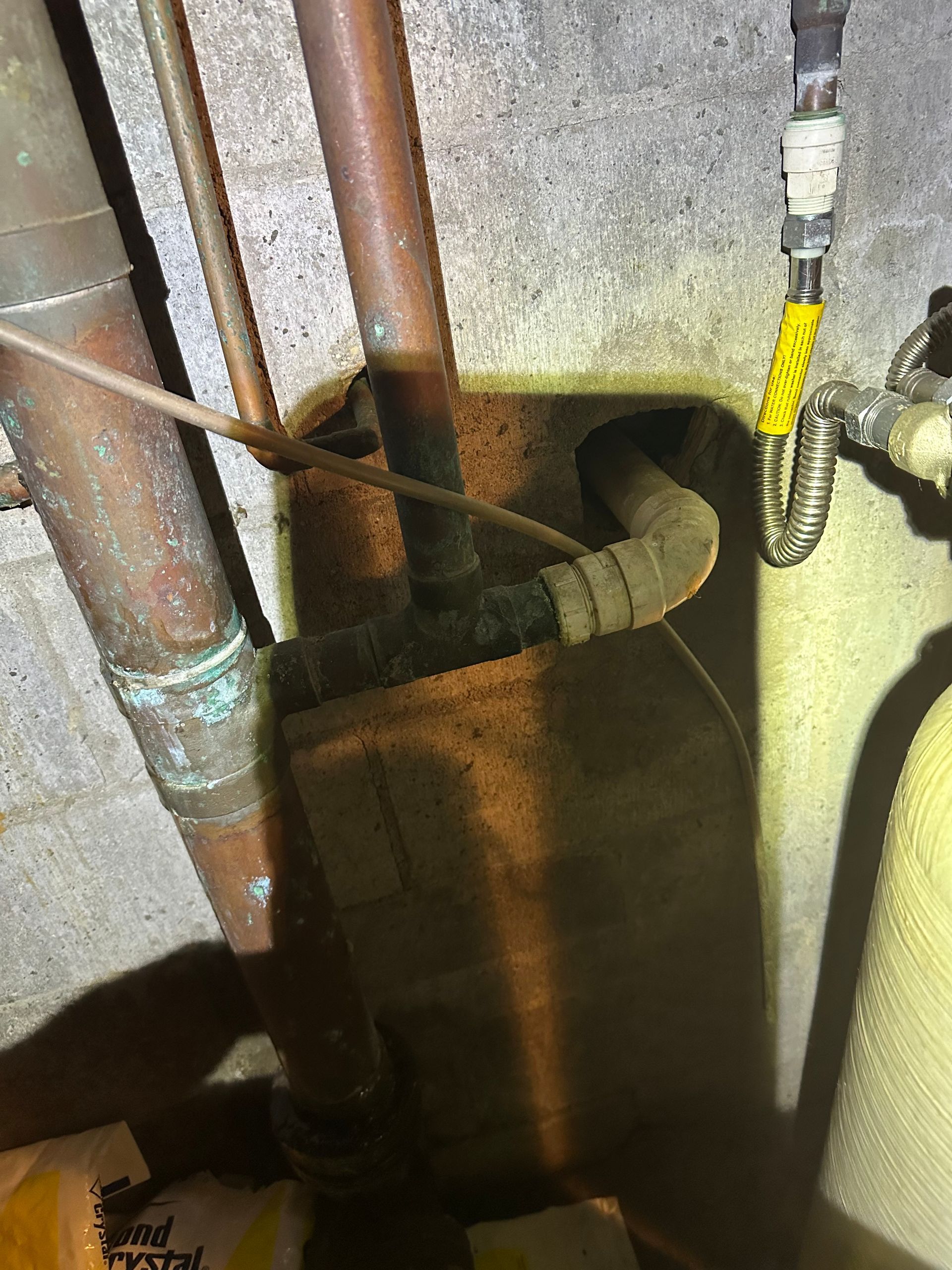 Damaged wall opening around pipes and a cable in a dim utility space