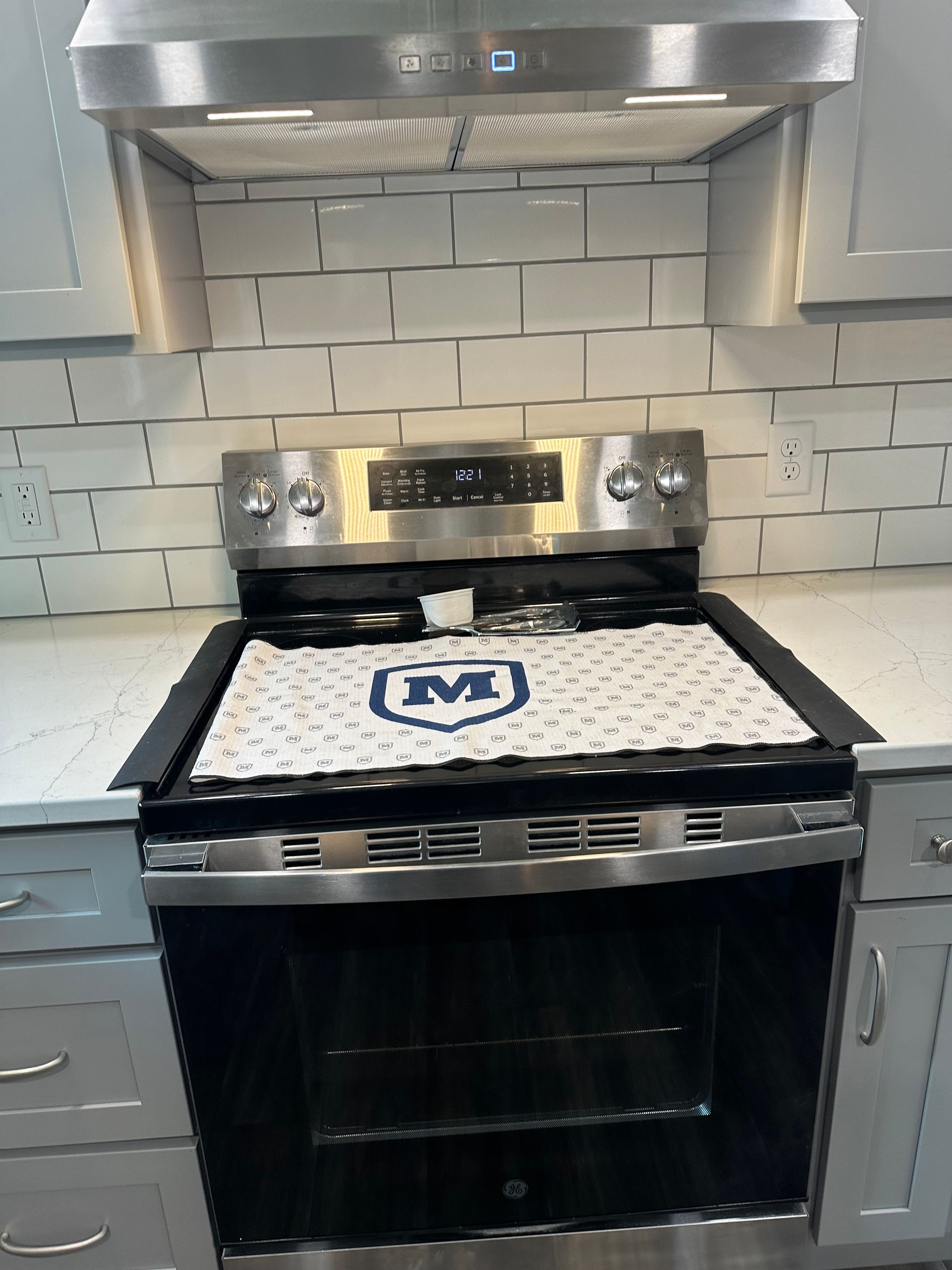 Kitchen stove with stainless steel range hood, white tile backsplash, and a white towel with a blue “M” on the oven handle