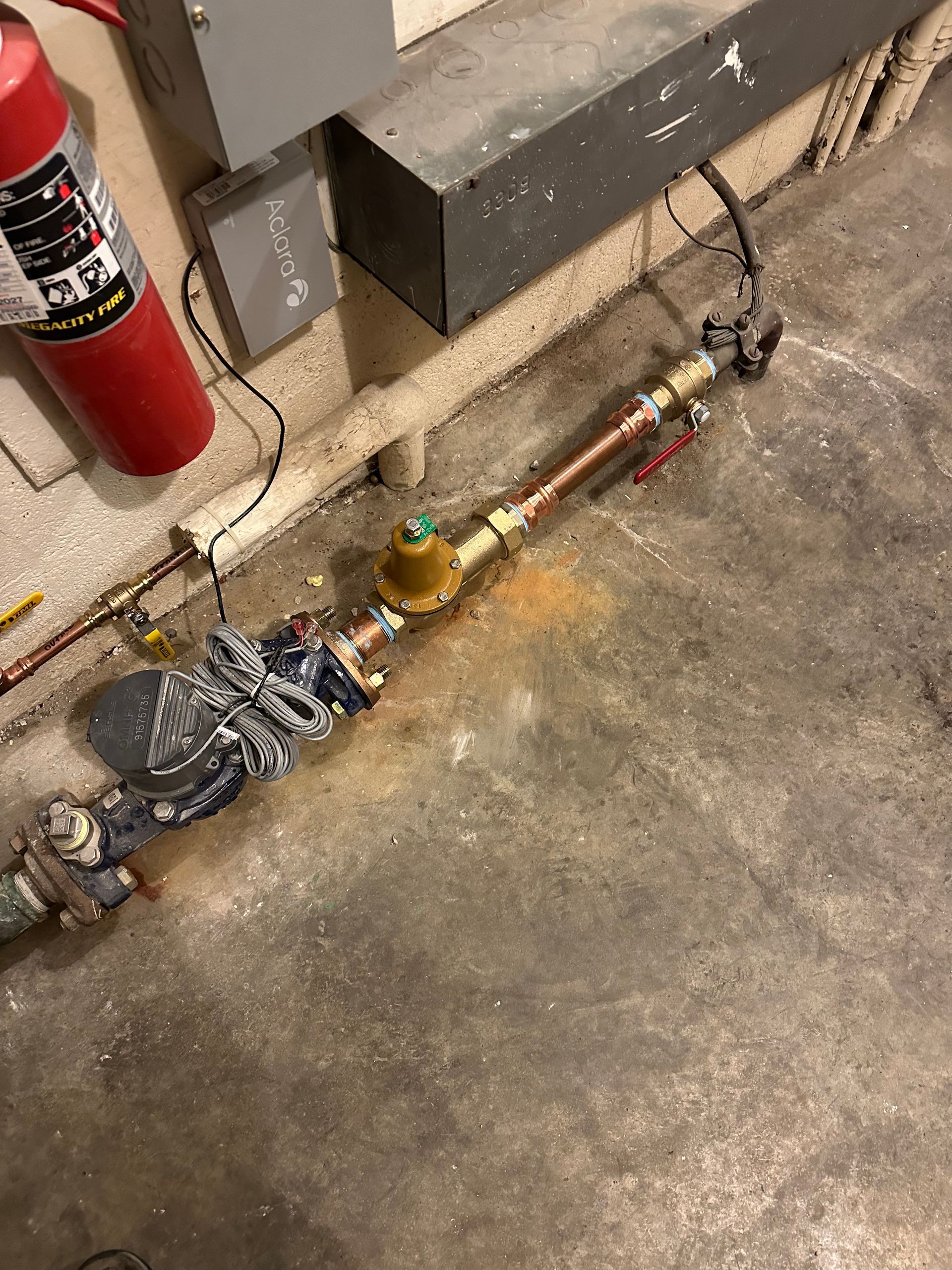 Fire extinguisher beside a floor-mounted valve assembly and pipe on a concrete floor