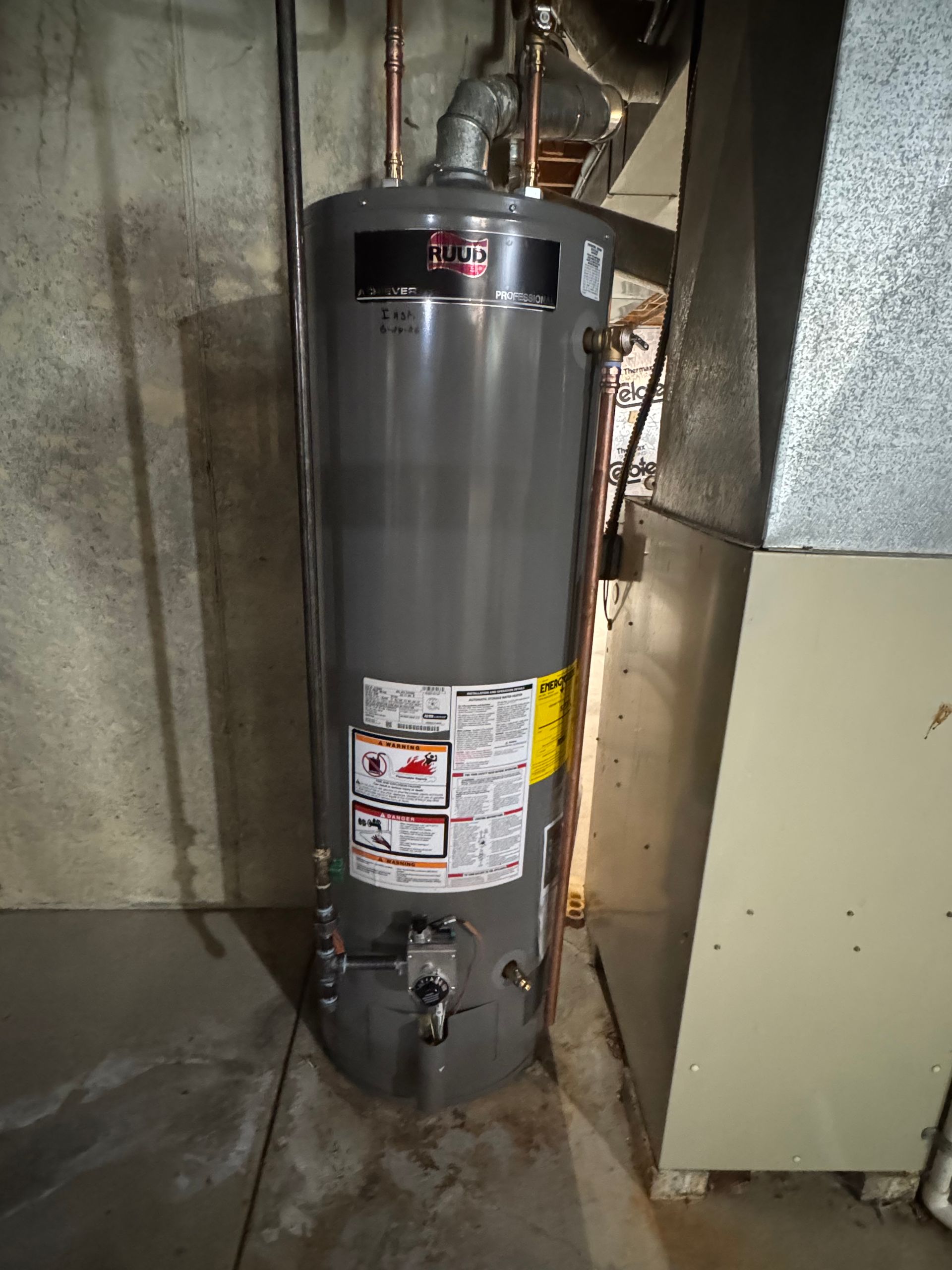 Gray water heater in a basement utility area with pipes and warning labels.