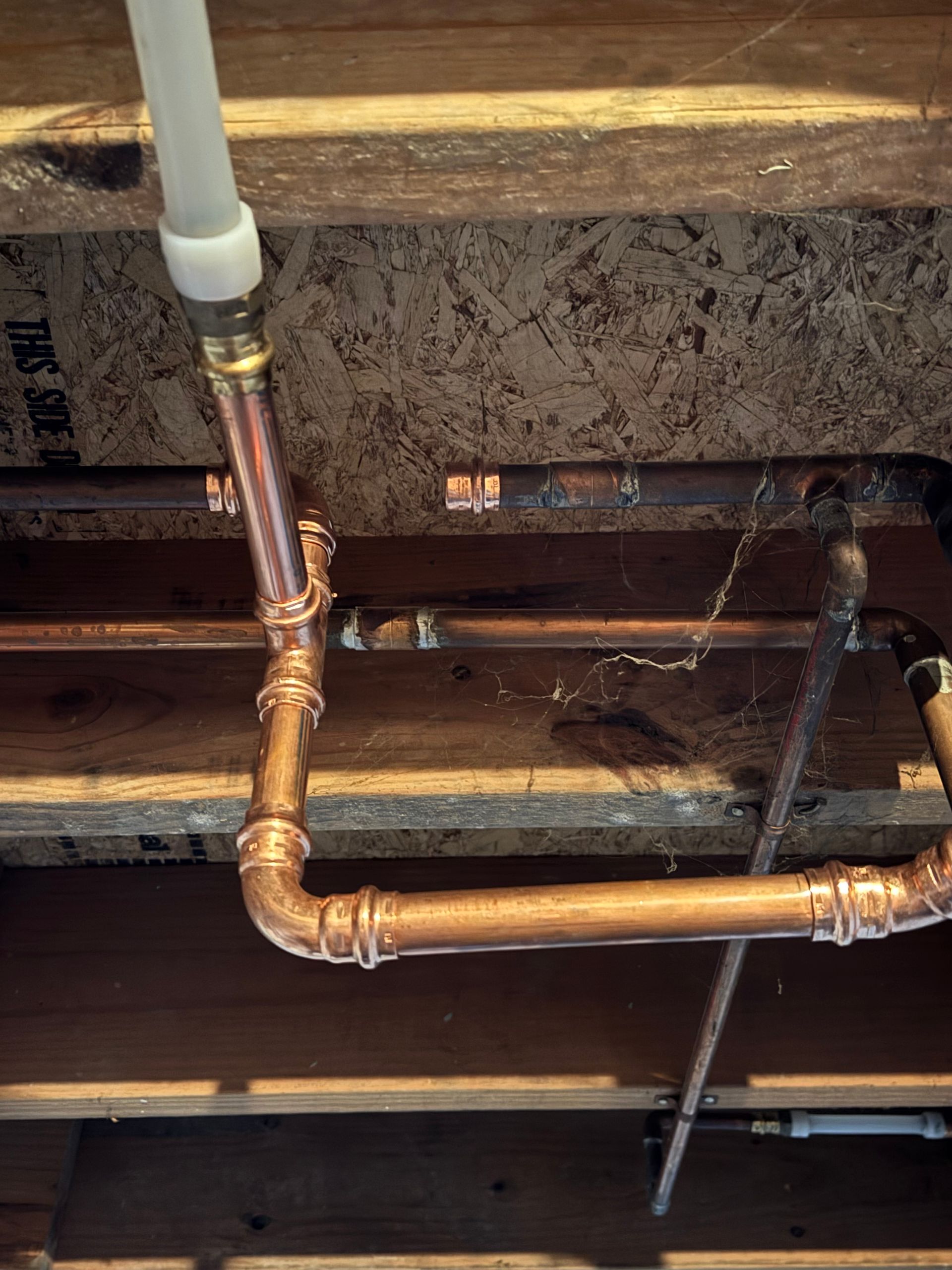 Copper plumbing pipes beneath wooden floor joists in a basement ceiling.