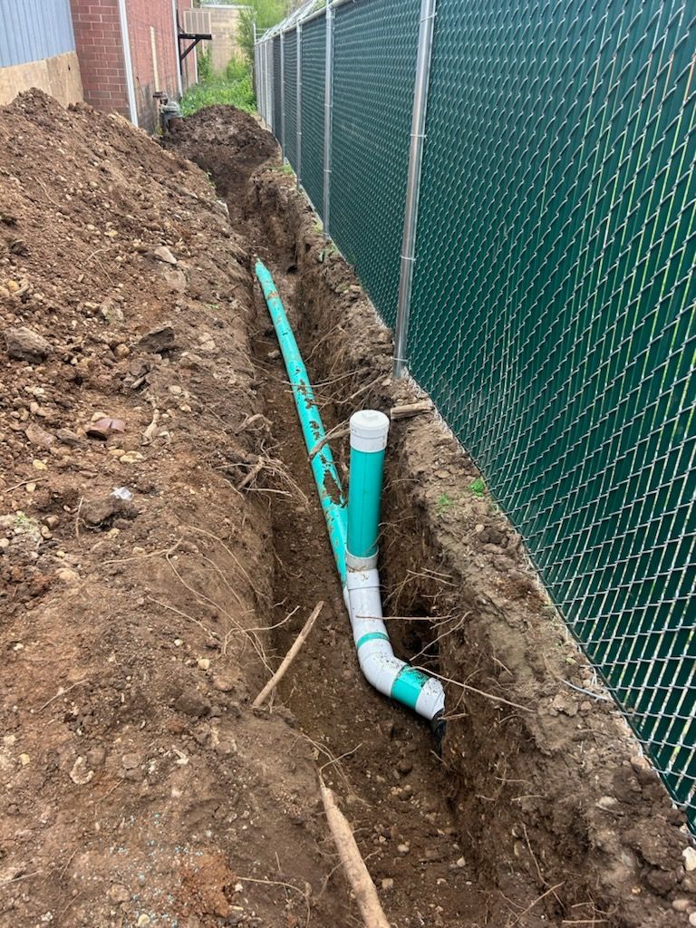 Trench beside chain-link fence with exposed green and white PVC pipes for utility work