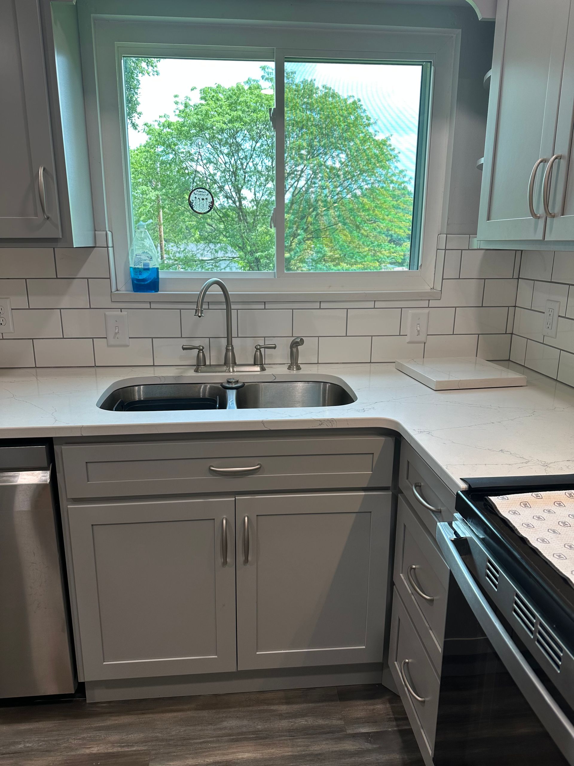 Bright kitchen sink beneath window with leafy trees outside, white cabinets and tile backsplash