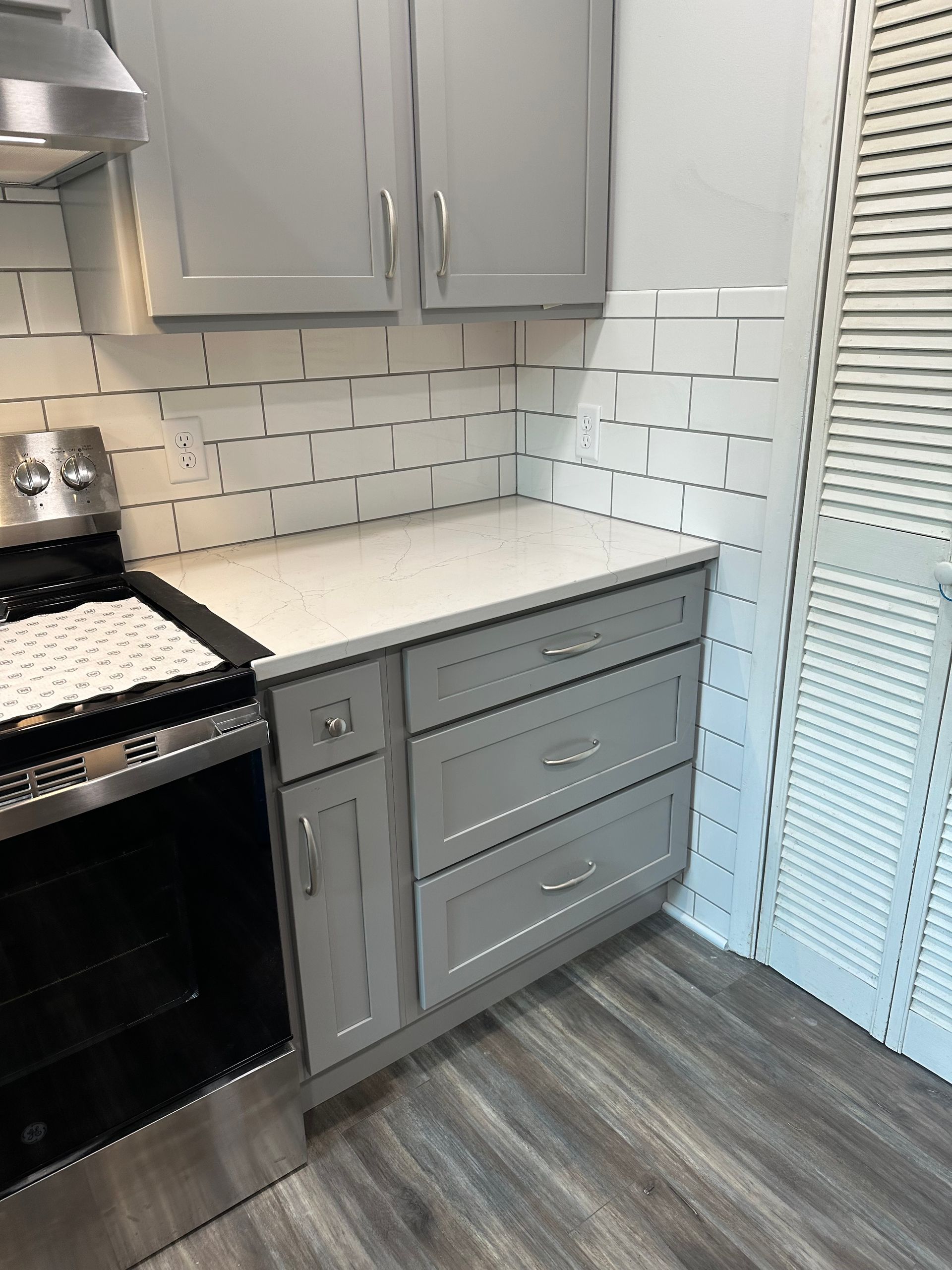 Small kitchen corner with gray cabinets, white backsplash, black stove, and louvered door.