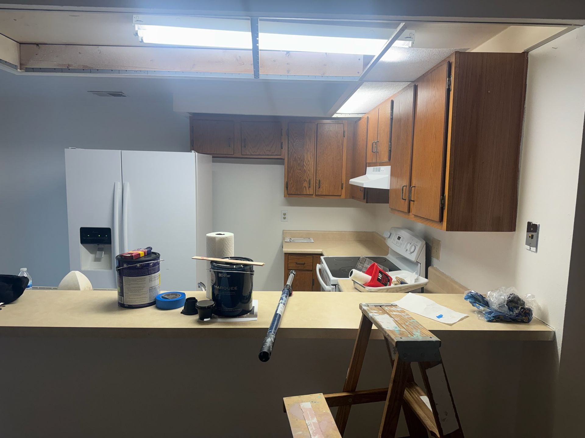Cluttered kitchen with white refrigerator, brown cabinets, countertop appliances, and a folded chair in the foreground