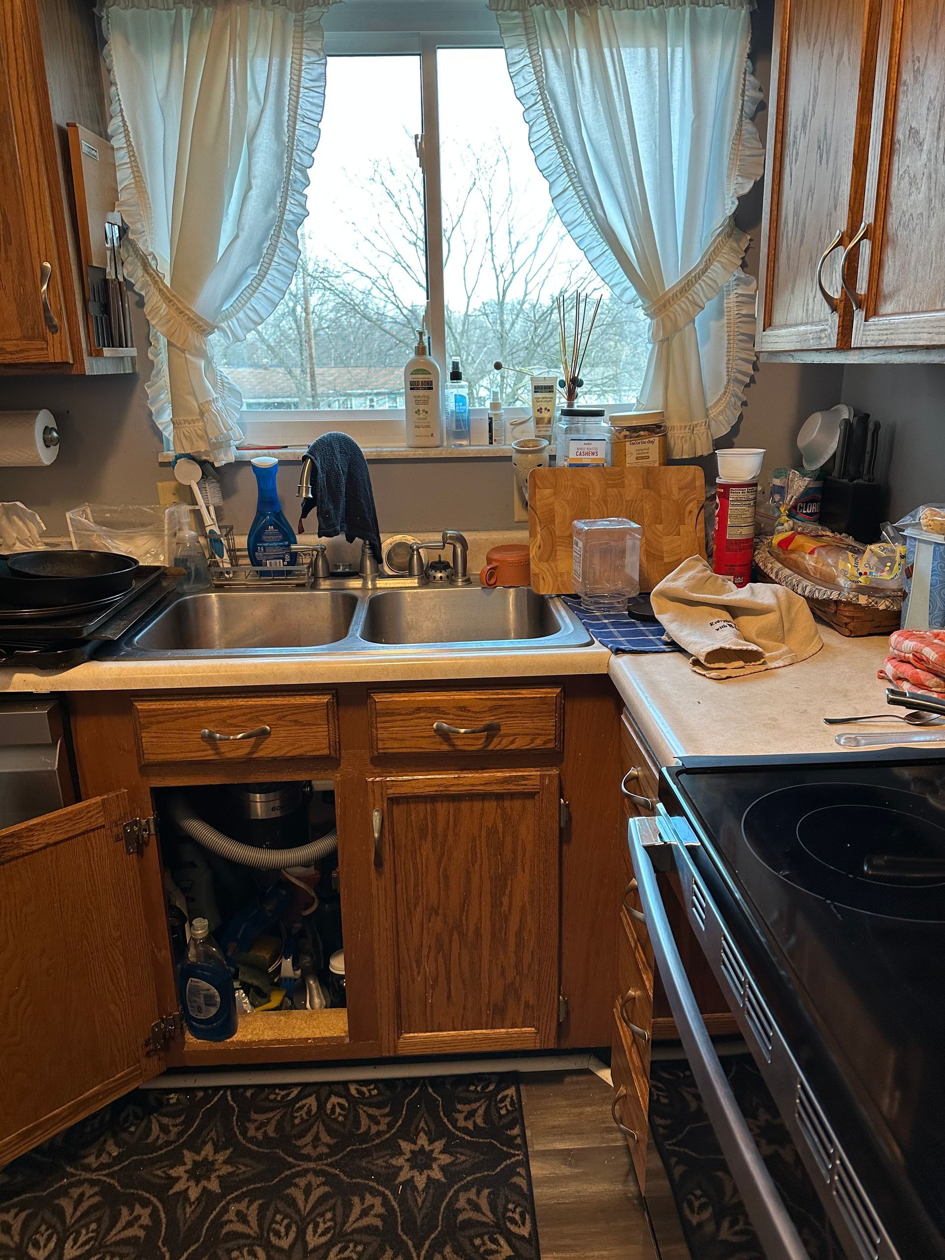 Small kitchen with wooden cabinets, sink under a curtained window, and a black stove beside a patterned rug.