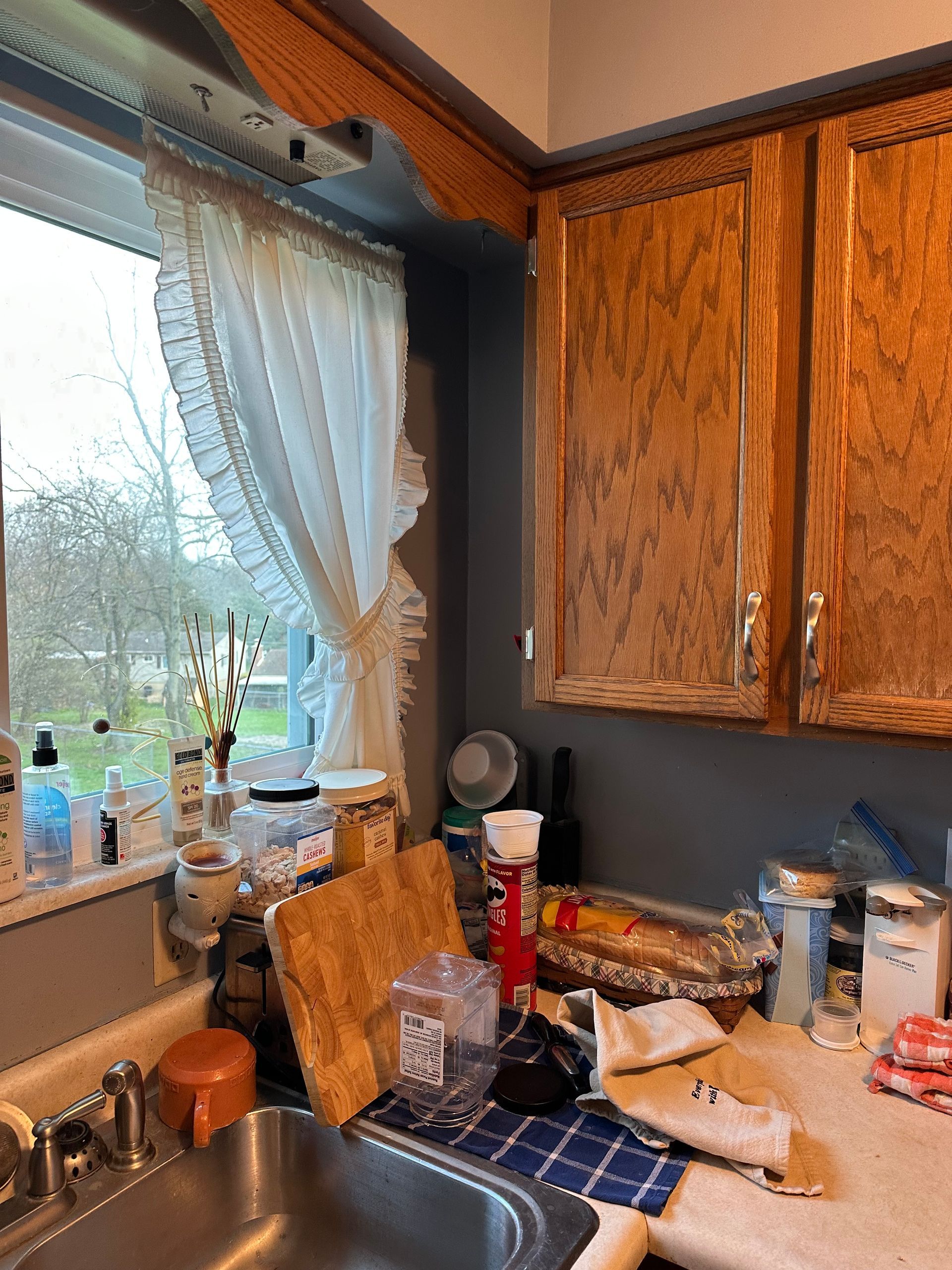Kitchen counter by a window with dishes, bottles, and wood cabinets, lit by daylight.
