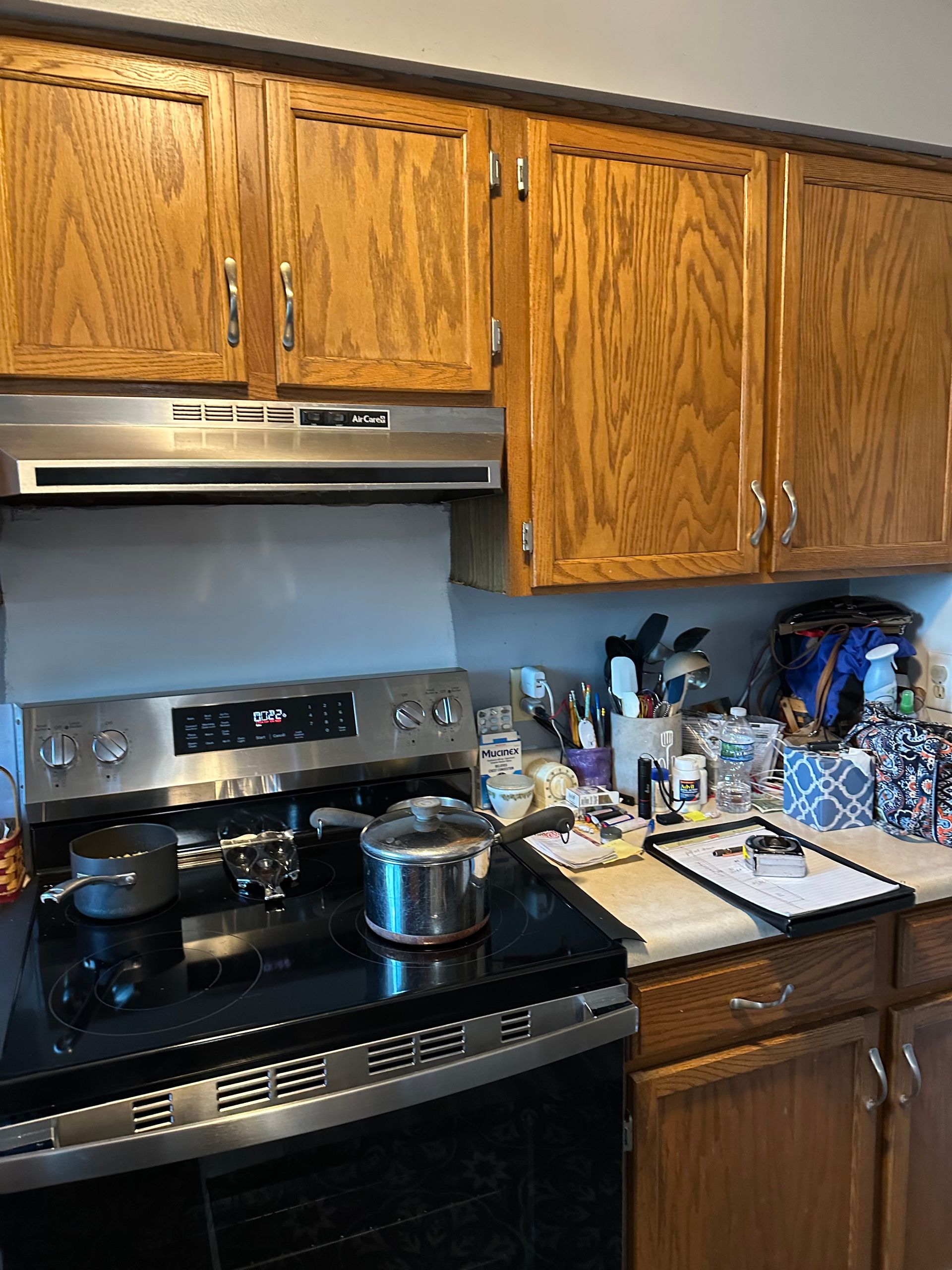 Kitchen stove and sink area with wood cabinets, a pot on the stove, and dishes cluttering the countertop.