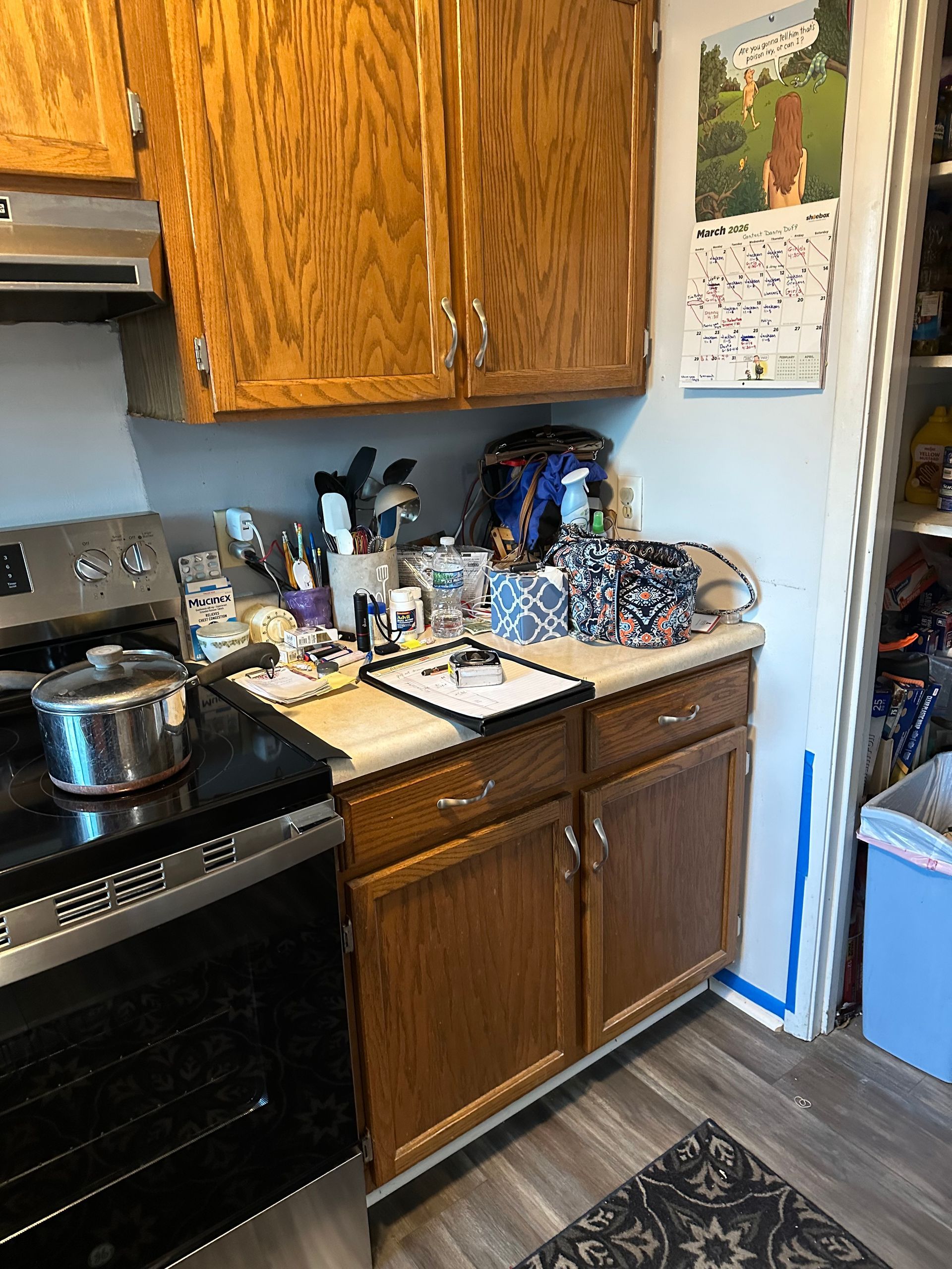 Cluttered kitchen counter beside a black stove, wooden cabinets, and a white wall calendar.
