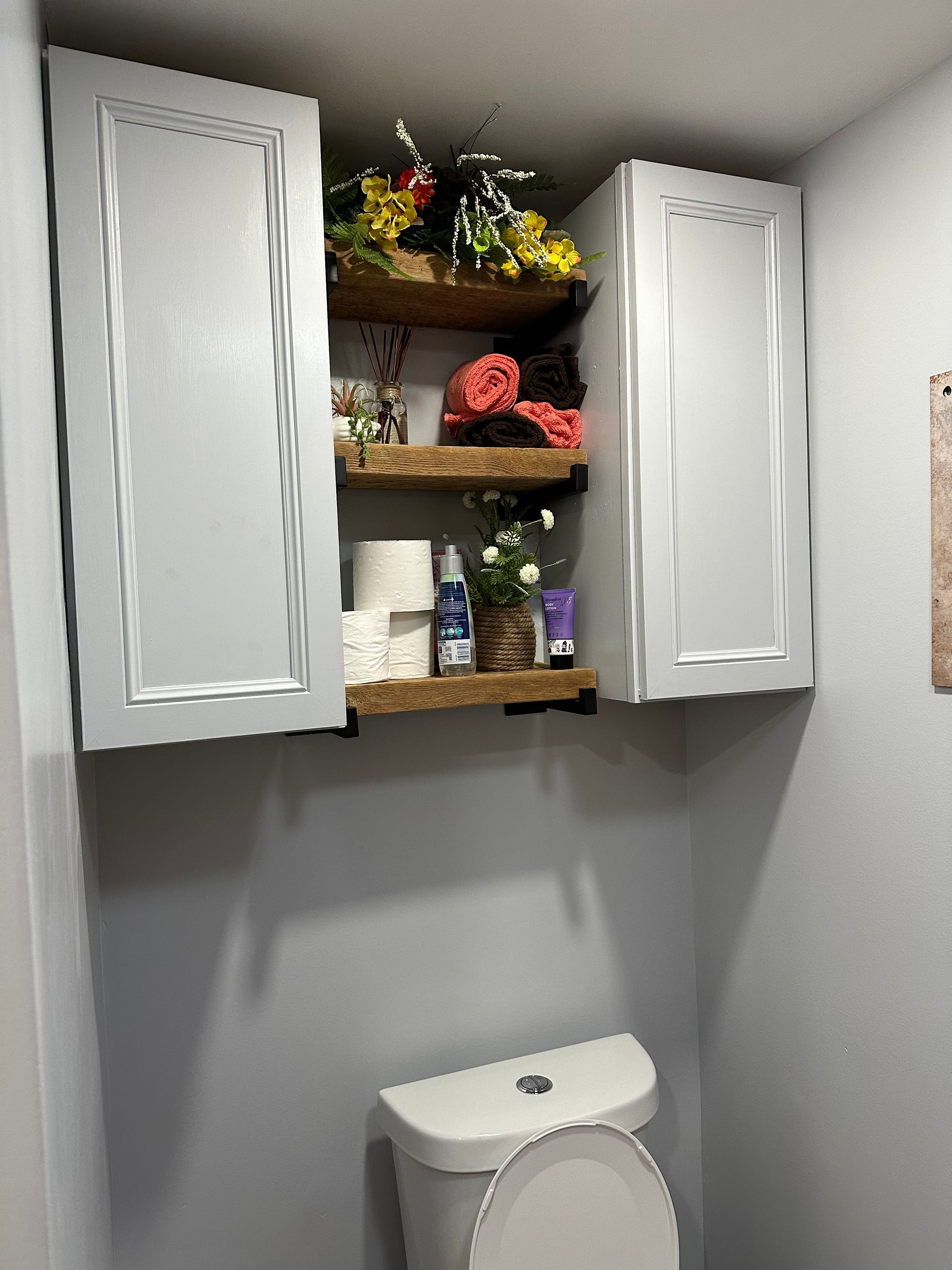 Small bathroom with gray walls, white cabinet shelves, flowers, and the top of a toilet below.