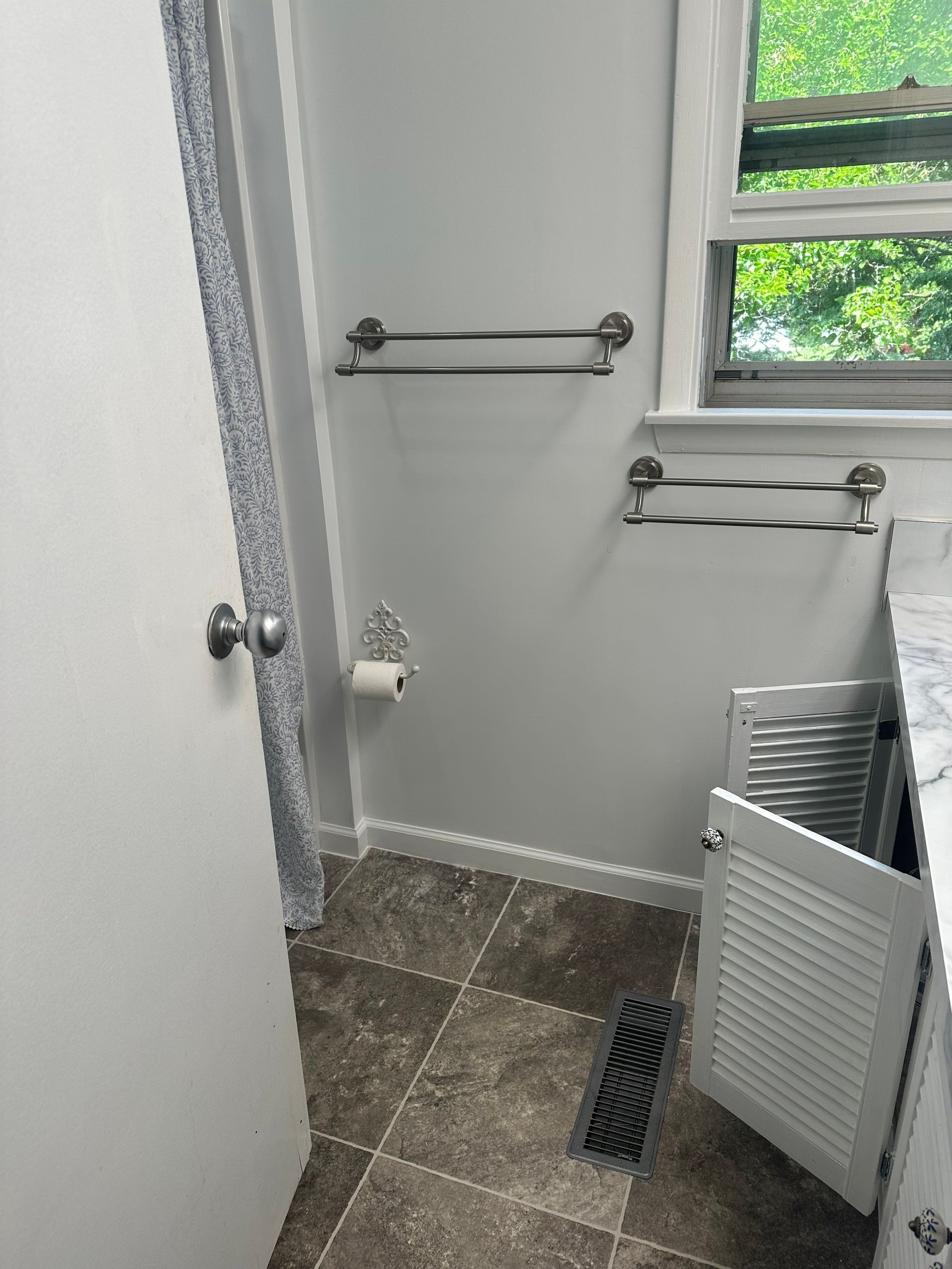 Small bathroom with gray walls, tiled floor, towel bars, toilet paper holder, and a window.
