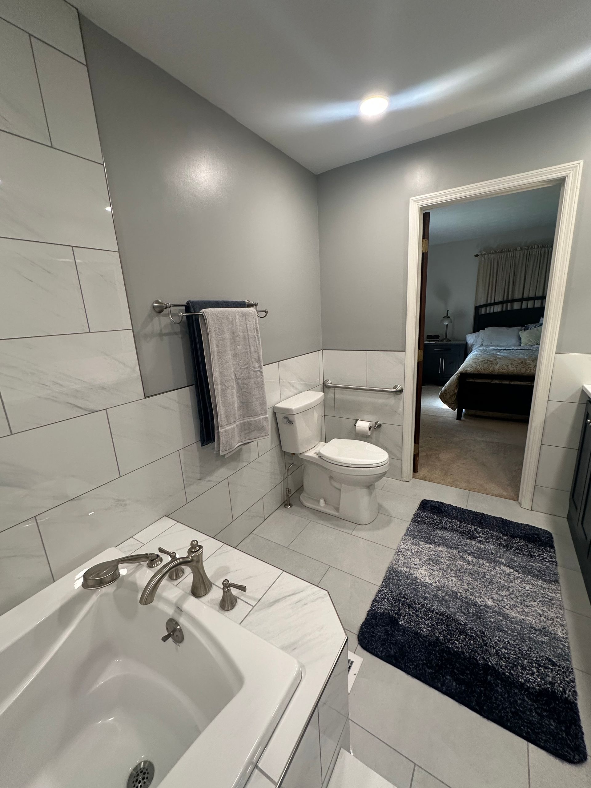 Bright bathroom with white tub, toilet, gray walls, blue rug, and doorway to a bedroom.