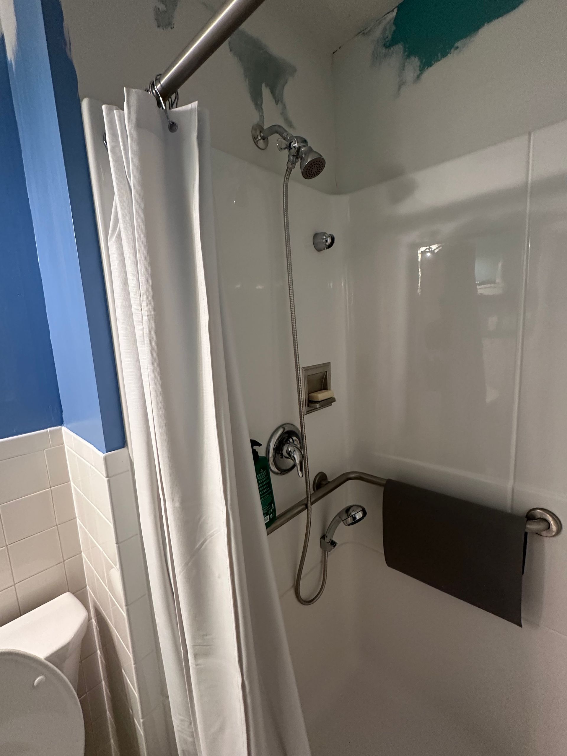 Shower stall with white curtain, grab bar, wall-mounted showerhead, and black seat in a tiled bathroom
