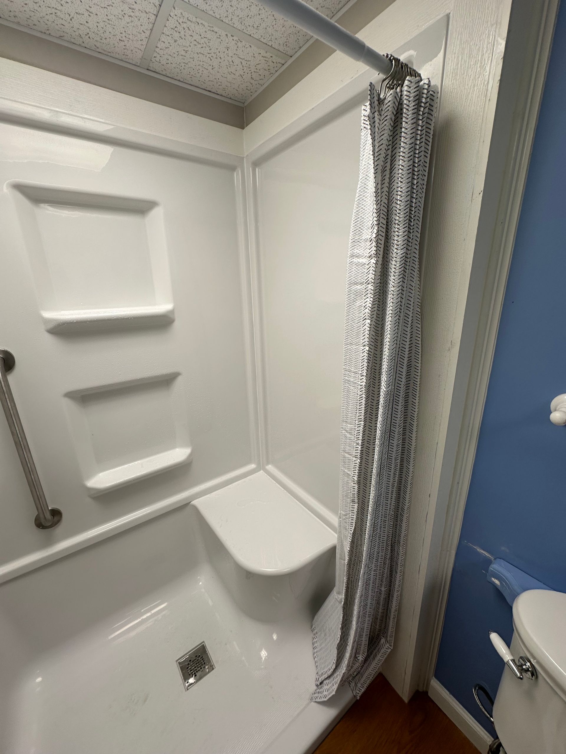 Small white shower stall with built-in seat, grab bar, and patterned shower curtain beside a blue wall.