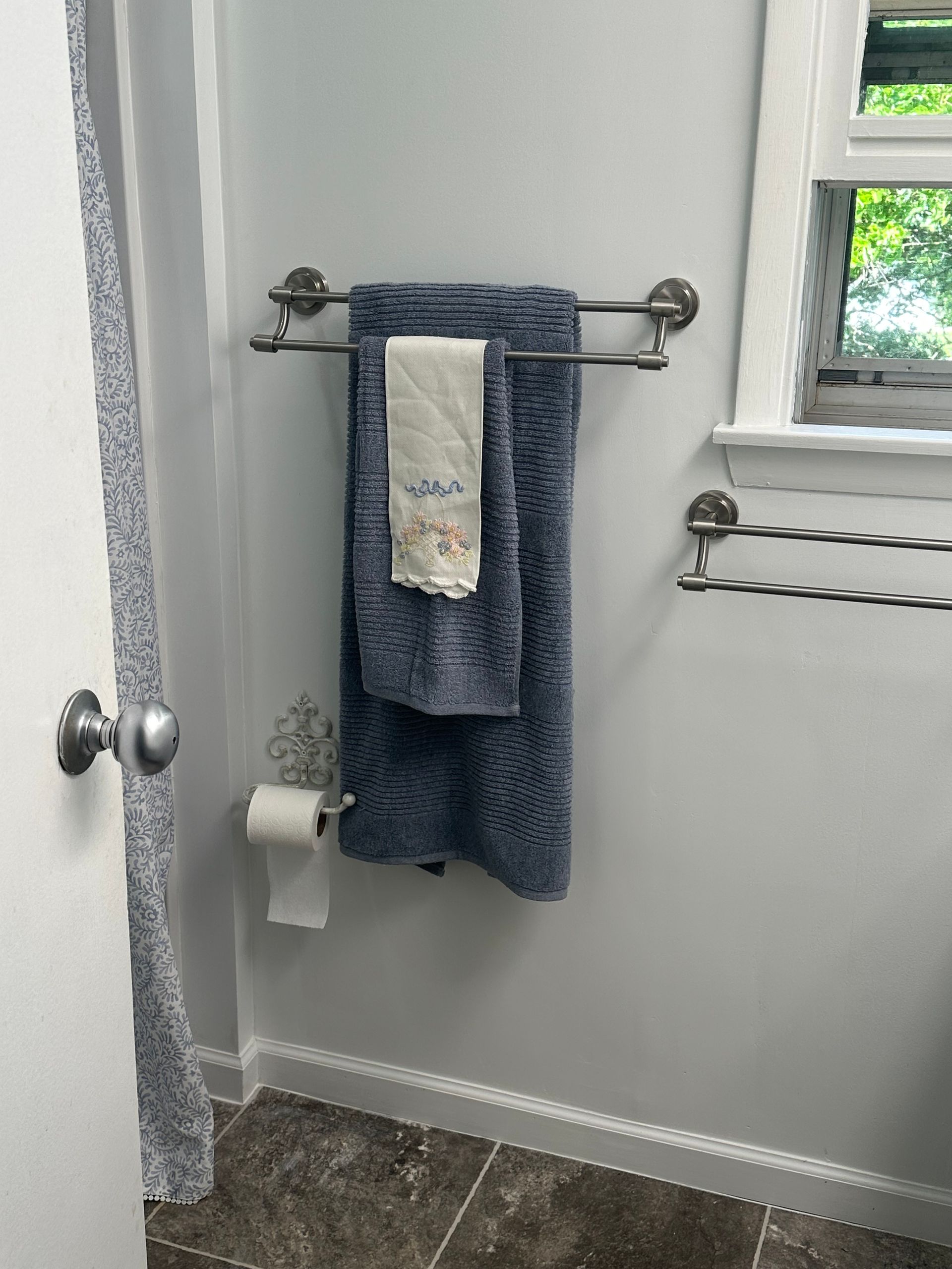 Bathroom towel rack with blue towels, toilet paper holder, and a window beside a shower curtain