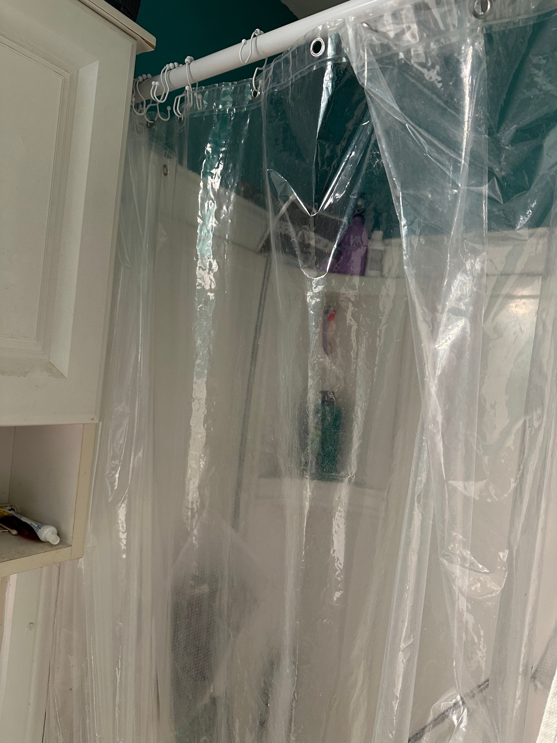 Clear plastic shower curtain hanging in a bathroom near a white wall and shelf.